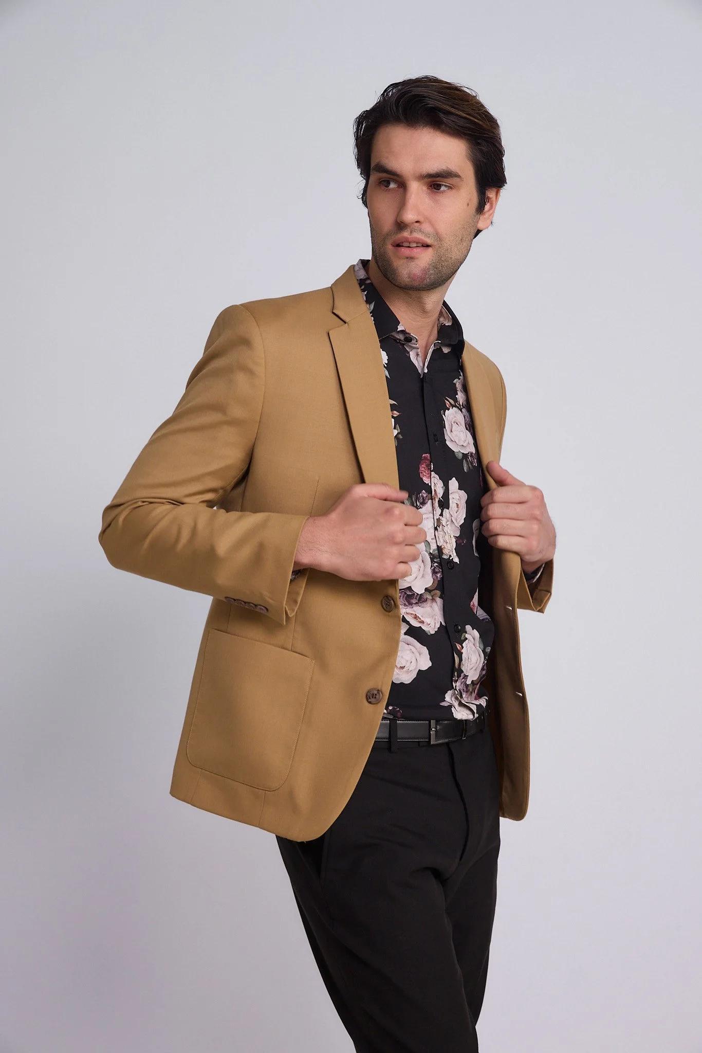 A man with dark hair wearing a tan blazer over a black floral shirt, adjusting his blazer against a plain light gray background.
