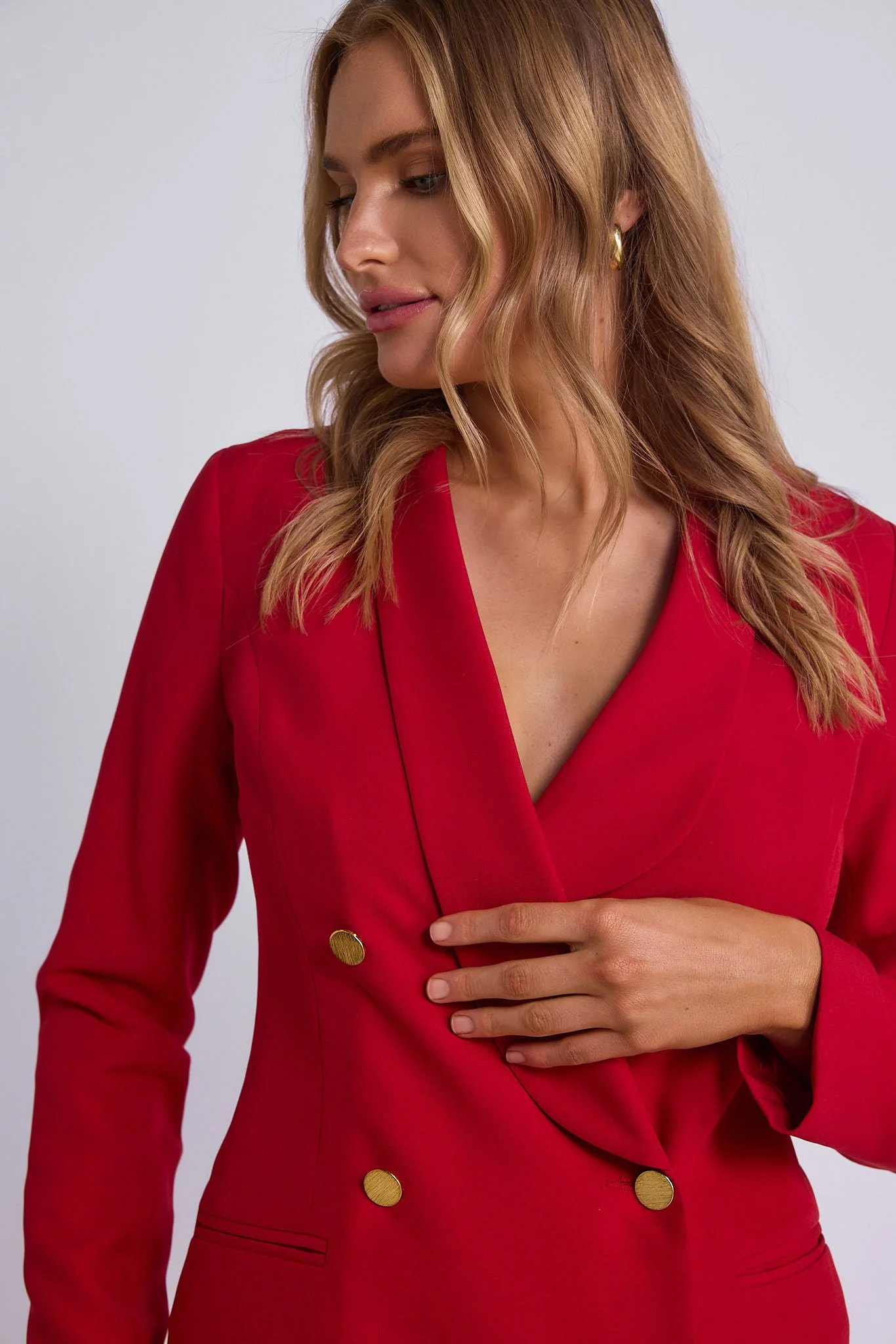 A woman with wavy blonde hair wearing a red blazer with gold buttons and gold hoop earrings, posing against a plain gray background.
