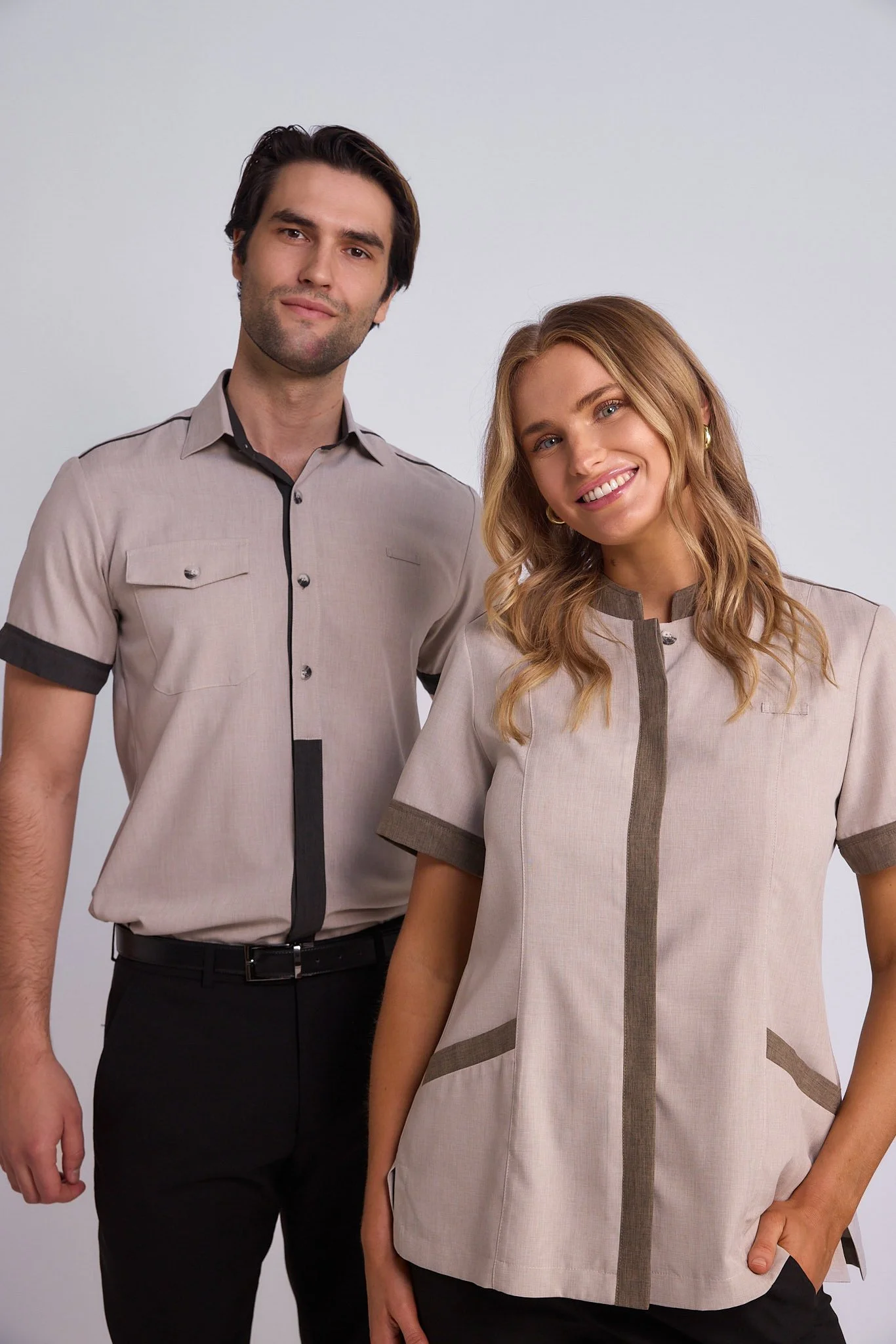 A man and a woman standing together indoors against a plain white wall, both wearing matching beige uniforms with dark accents.