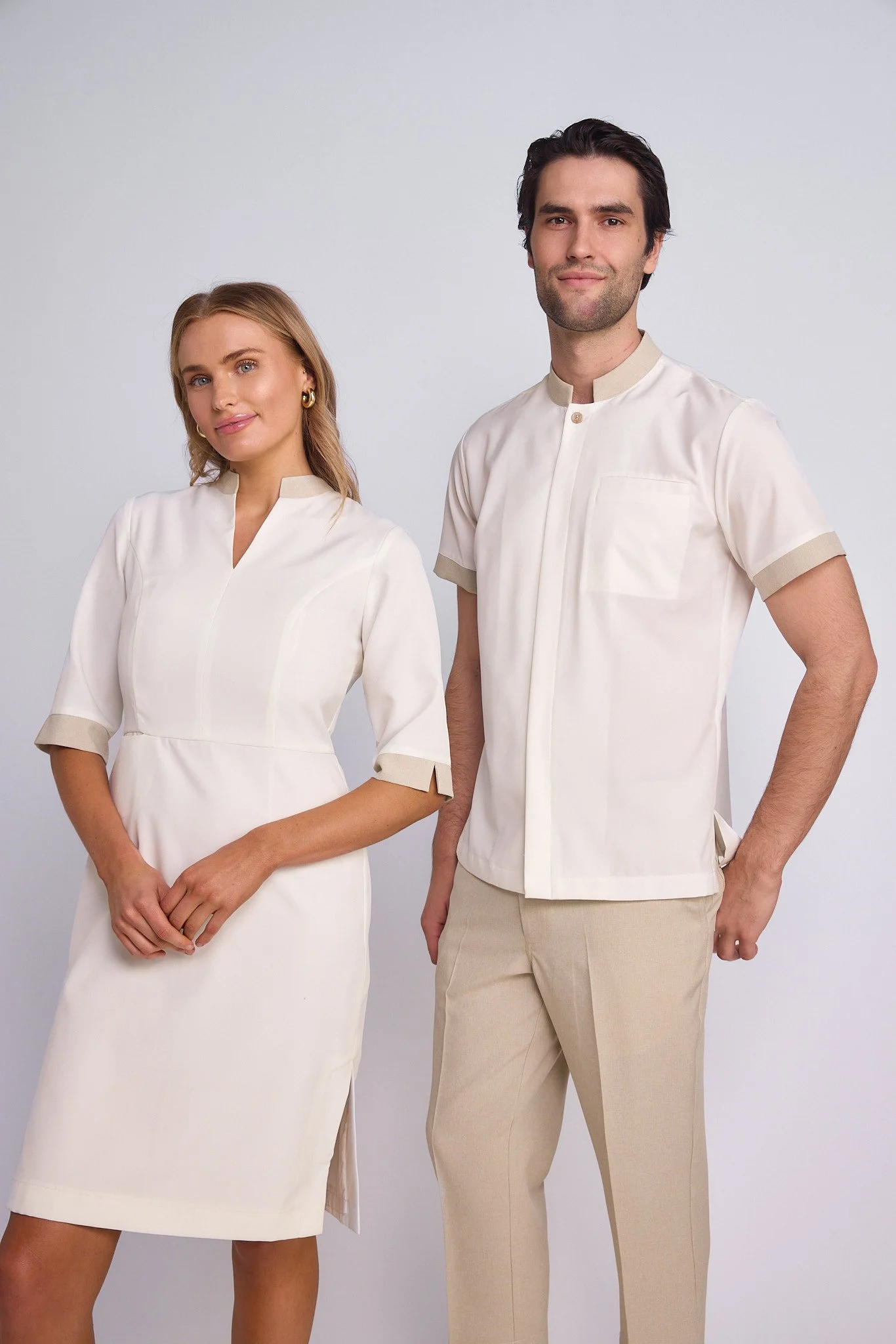 Two models, a woman and a man, standing against a plain white background, wearing matching cream-colored outfits. The woman is dressed in a cream dress with beige accents on the sleeves, and the man is wearing a cream short-sleeved shirt with beige a
