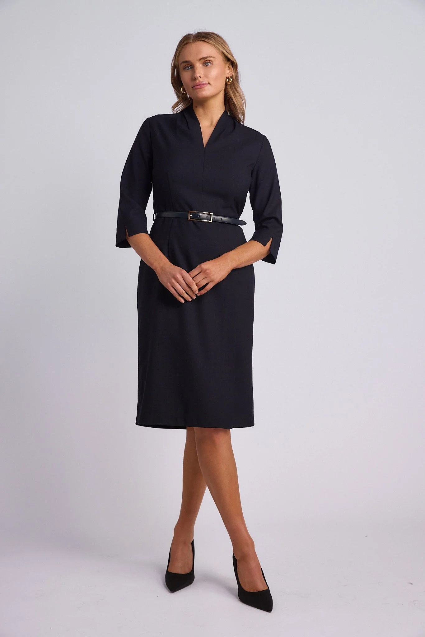 A woman wearing a black dress with three-quarter sleeves and a belt, standing against a plain background.