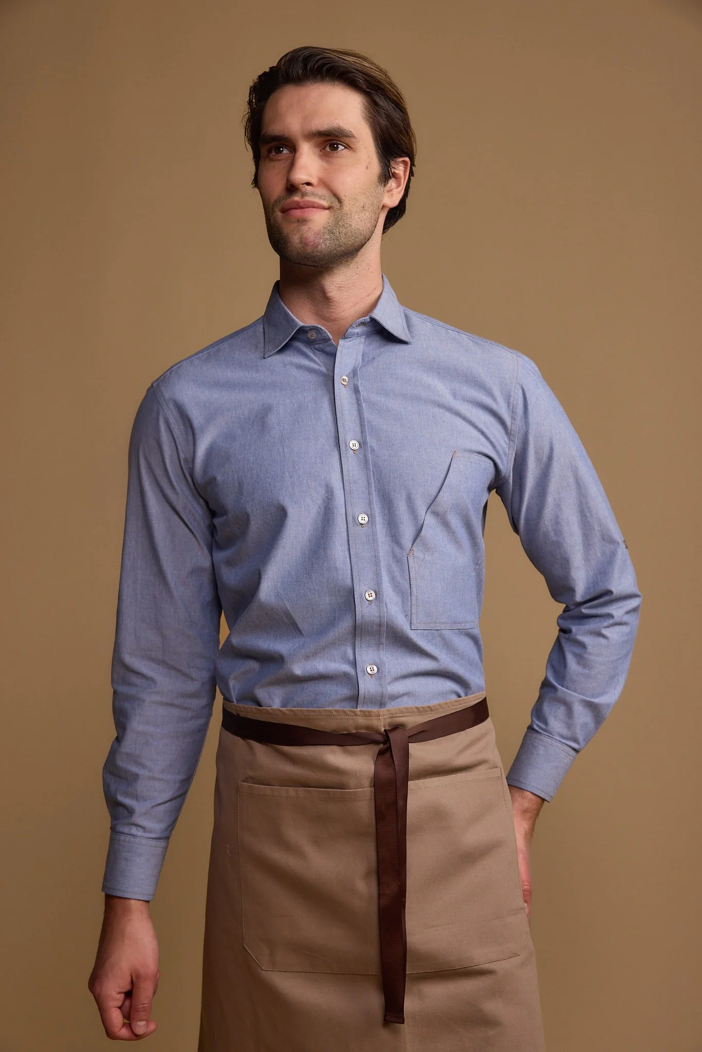 Man wearing a blue button-up shirt and a tan apron with a brown tie, standing against a plain beige background.