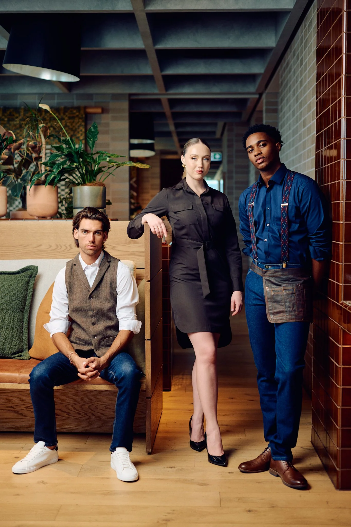 Three people in a modern restaurant interior, a man sitting on a bench, a woman standing with her hand on a divider, and a man standing with his hands behind his back. The setting features wooden flooring and decorative plants.