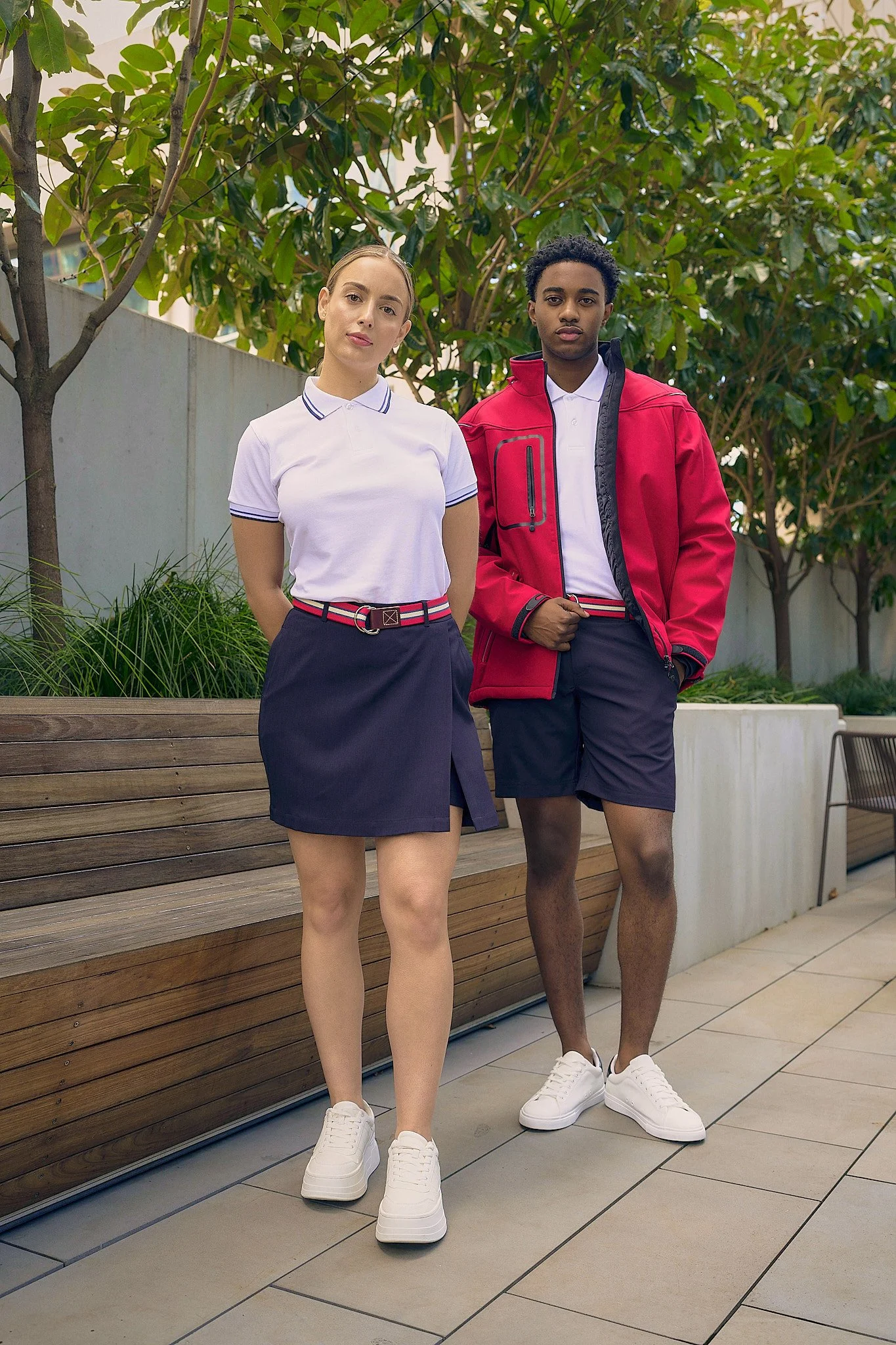 A young woman and a young man standing outdoors on a tiled patio, surrounded by green trees and bushes. They are dressed in casual clothing with sporty and preppy styles, both wearing white sneakers.
