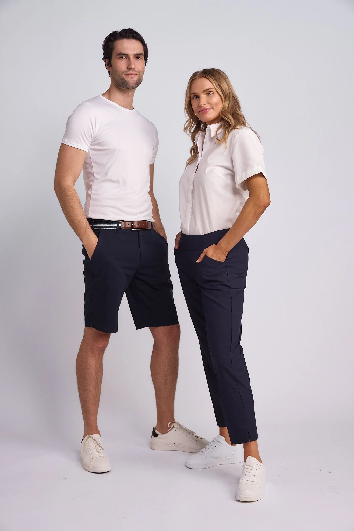 A man and woman standing side by side against a plain light gray background. The man is wearing a white t-shirt, dark shorts, and white sneakers. The woman is wearing a white short-sleeve blouse, dark pants, and white sneakers. Both have their hands 