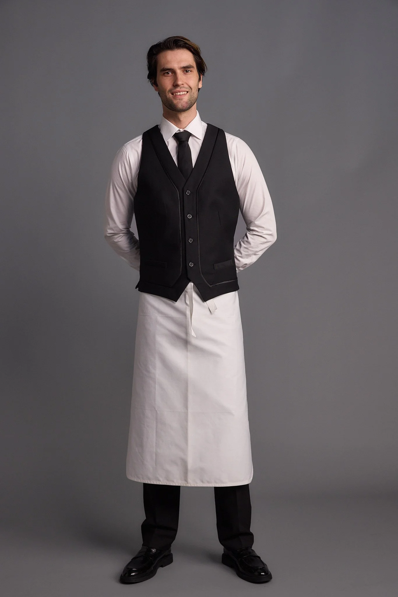A man dressed as a waiter wearing a white shirt, black vest, black tie, long white apron, black trousers, and black shoes, standing against a gray background.