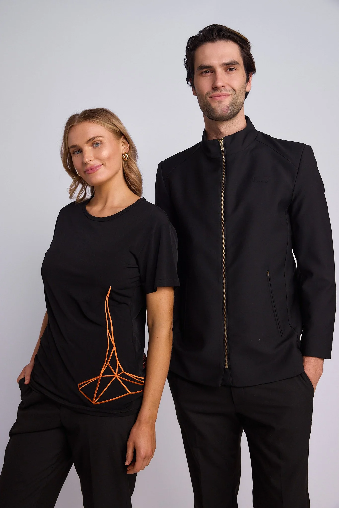 A man and woman standing side by side against a light gray background, both wearing black clothing. The woman has shoulder-length blonde hair and is smiling slightly, wearing a black t-shirt with an orange geometric pattern on the lower left side. Th