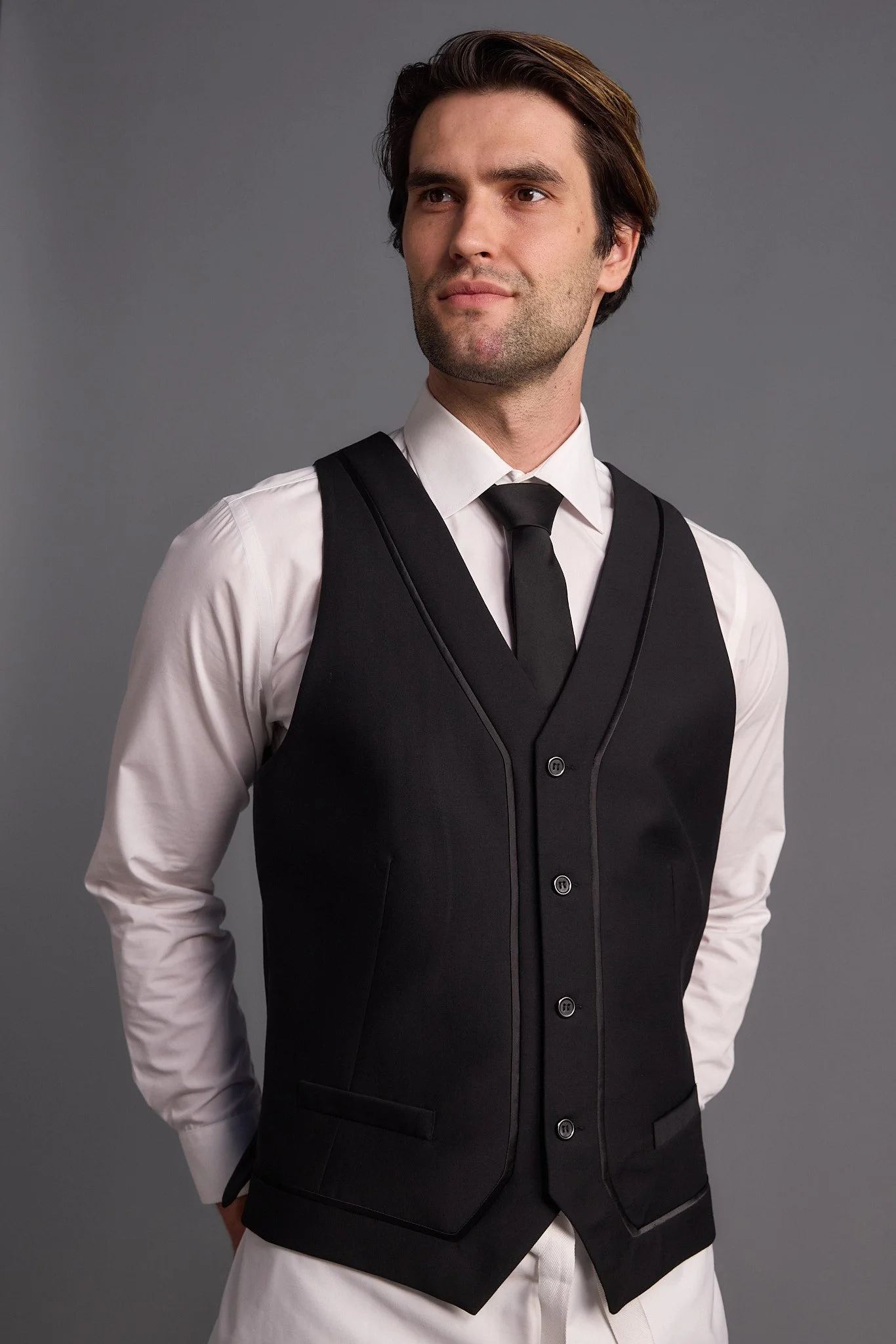 A man in a white dress shirt, black vest, black tie, and white pants standing against a plain gray background.