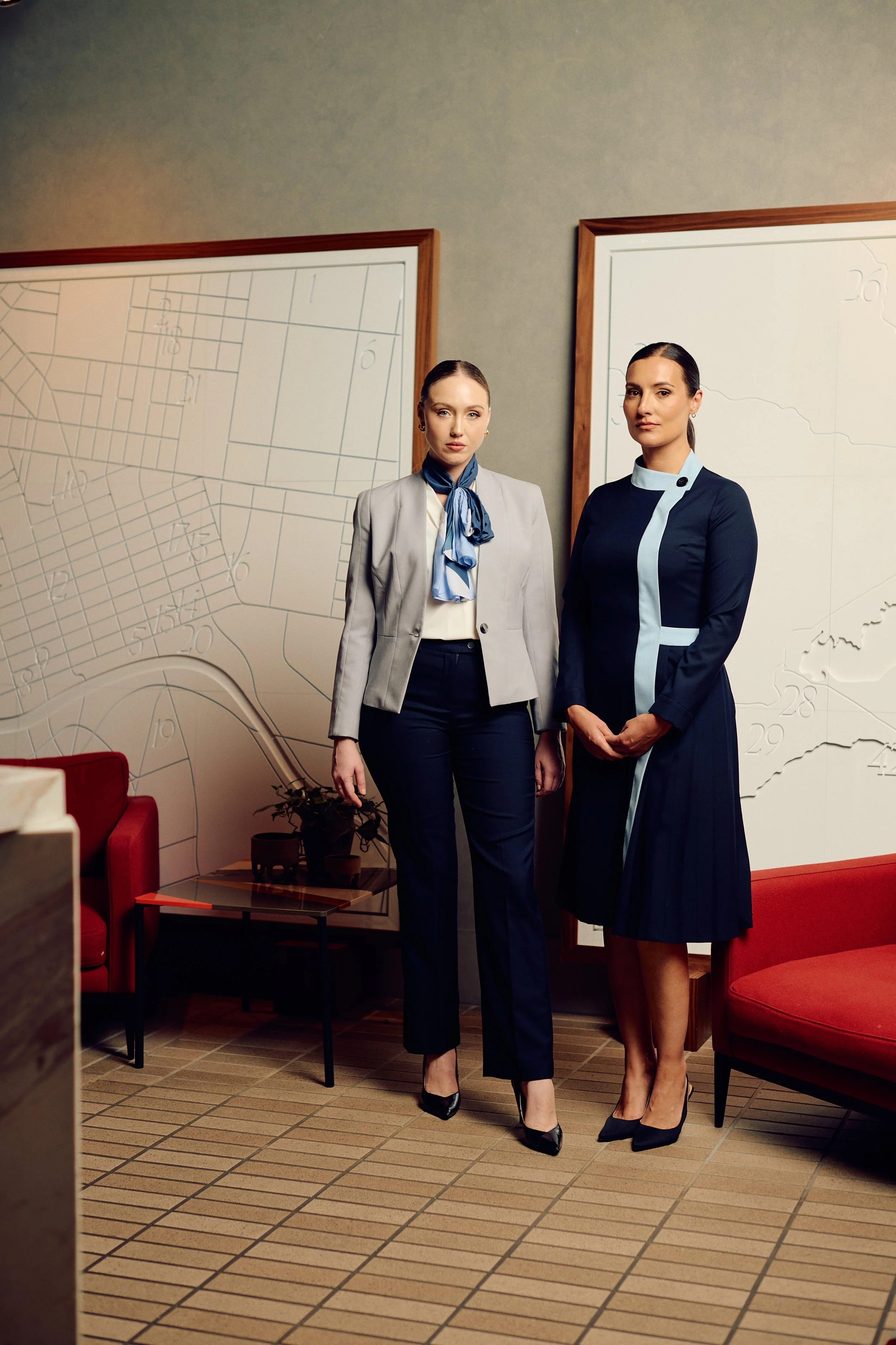 Two women standing side by side in a modern office or conference room, with large whiteboards featuring diagrams or maps behind them, and red seating nearby.