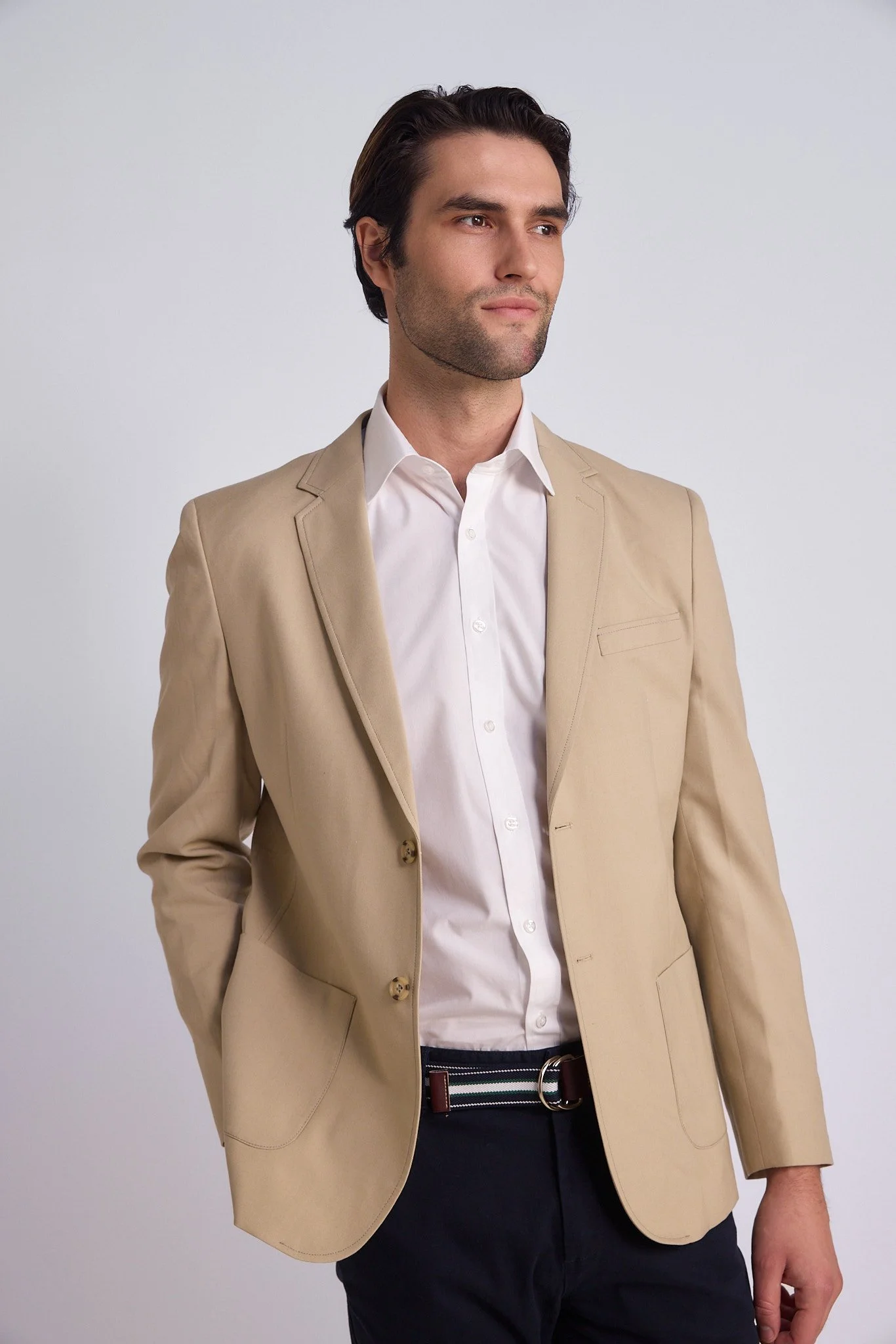 A man in a beige suit jacket, white dress shirt, and black pants standing against a neutral background.
