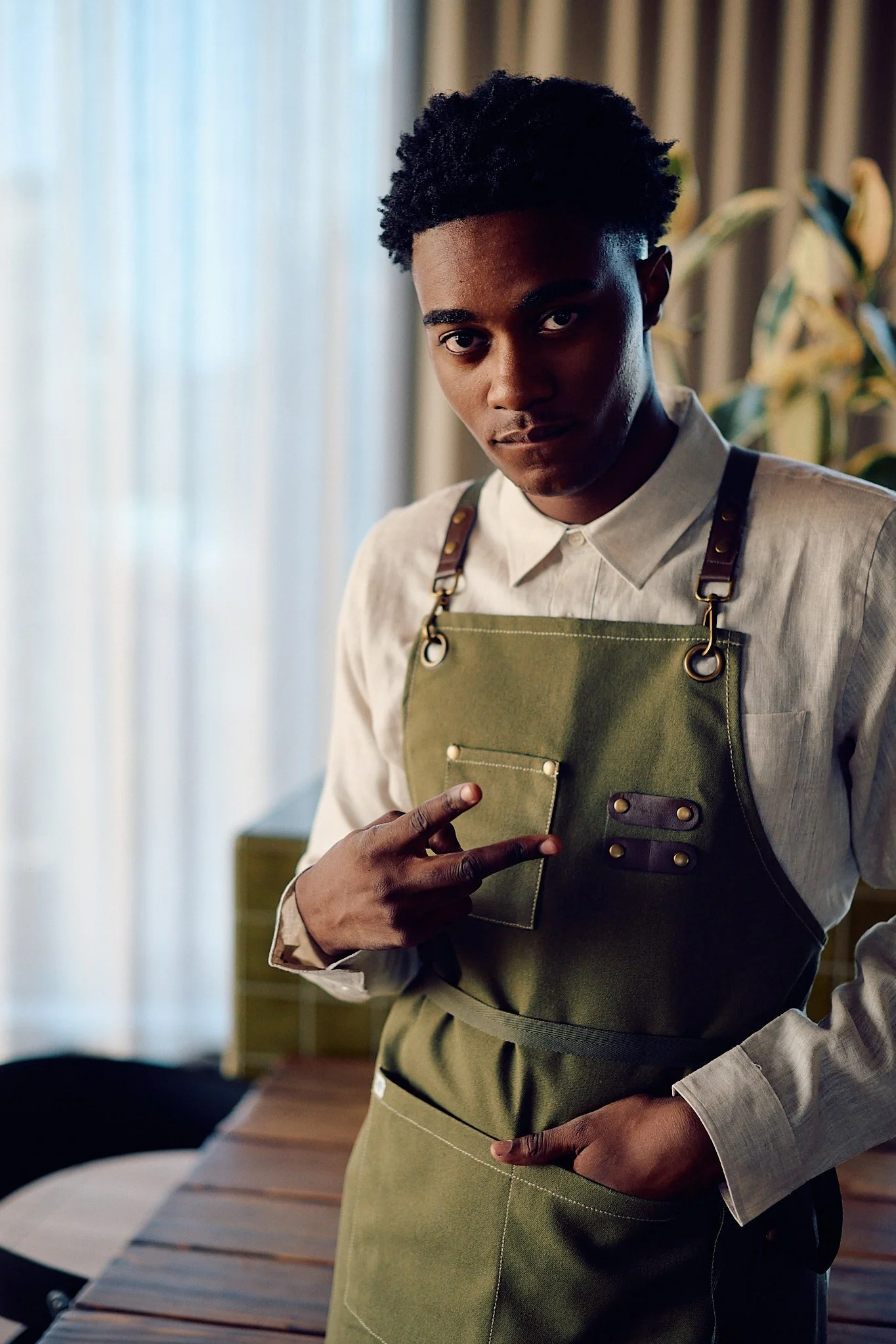 Young man wearing a beige shirt and green apron with leather straps, standing indoors near a window with curtains, pointing to himself.