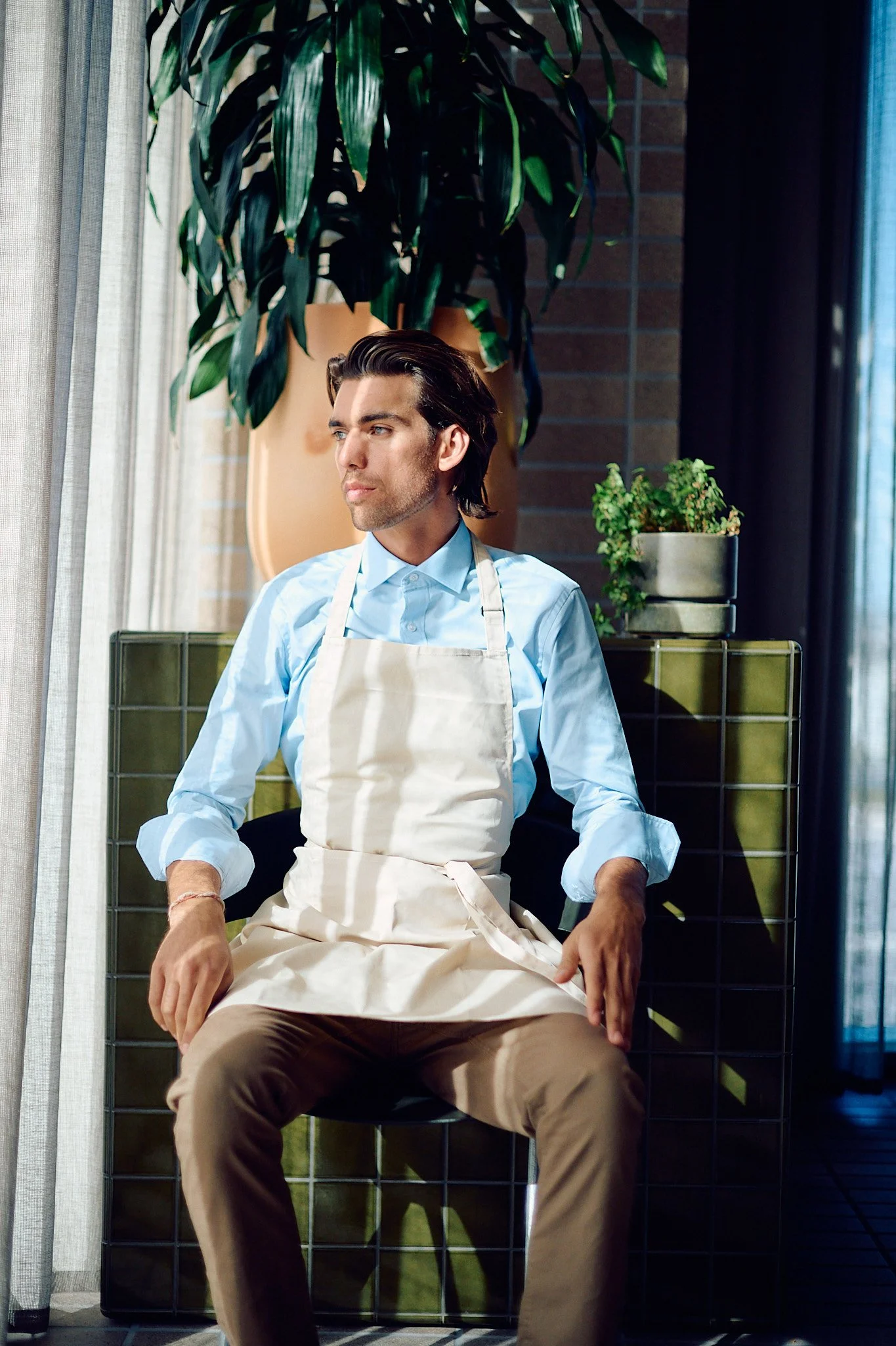 A man with dark hair, wearing a light blue shirt and beige apron, sitting on a black chair indoors, with large green potted plants behind him and natural light coming through a window.