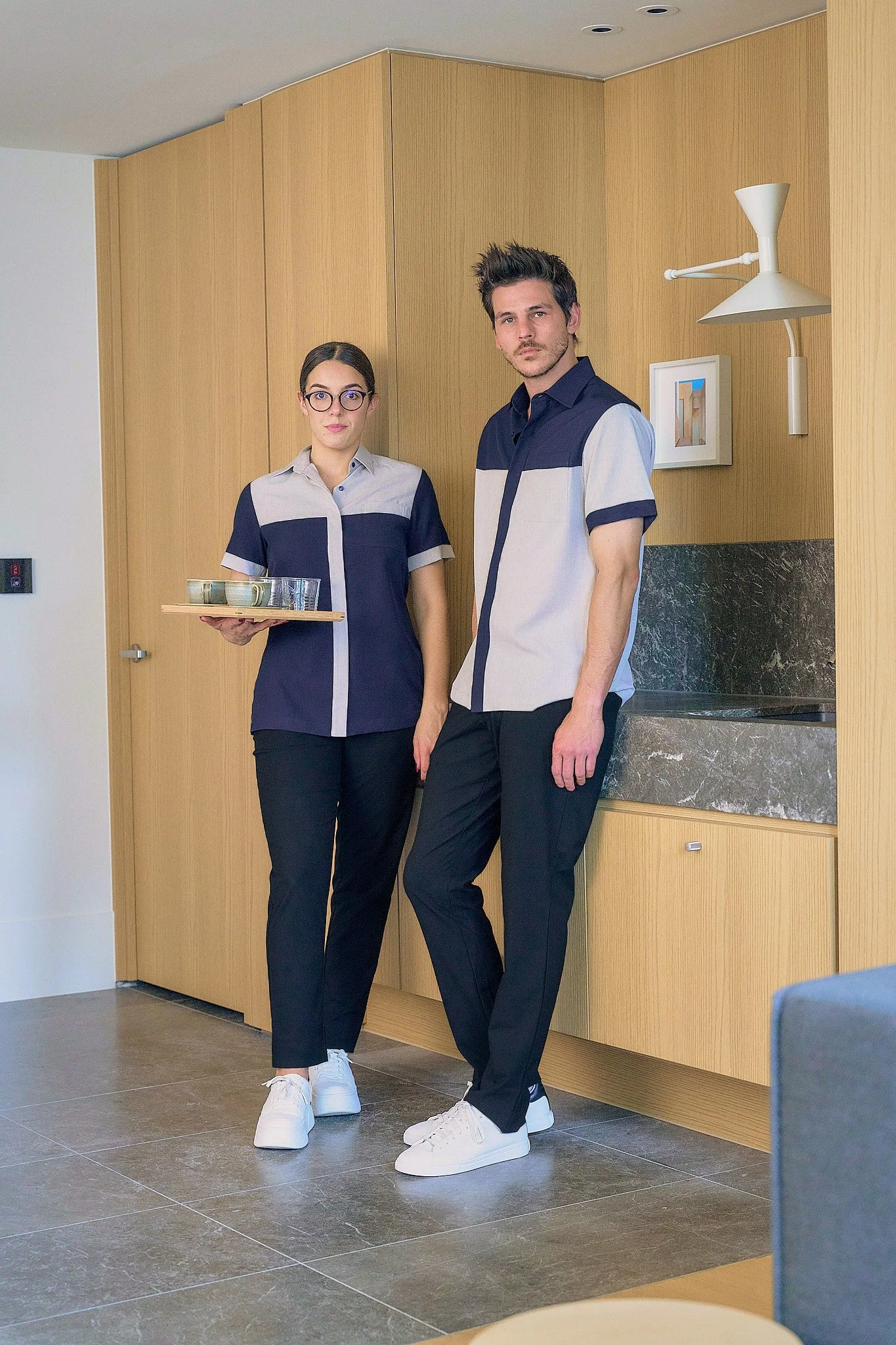 Two hotel staff members, a woman and a man, standing against a wooden wall, with the woman holding a tray of drinks. They are dressed in coordinated uniforms with navy and grey shirts, black pants, and white sneakers.