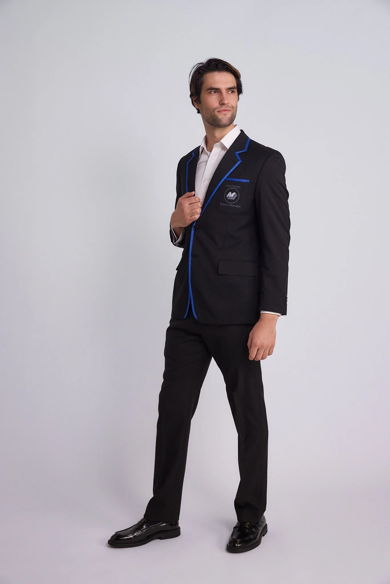 A man in a black suit with blue trim, white shirt, and black shoes, standing against a plain white background, holding the lapel of his suit jacket.