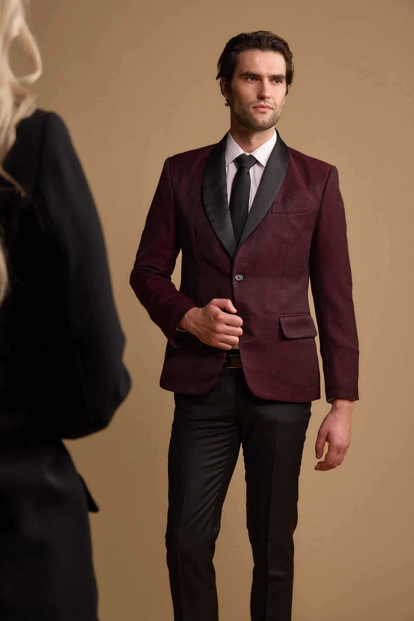 A man in a burgundy blazer with black satin lapel, white shirt, and black tie, standing against a beige background.