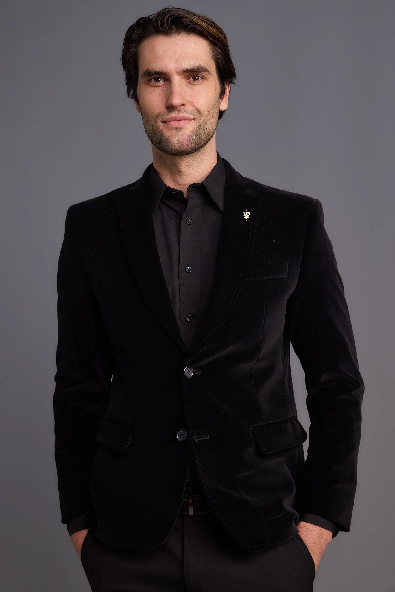 A man with dark hair, wearing a black suit jacket and black shirt, stands against a gray background