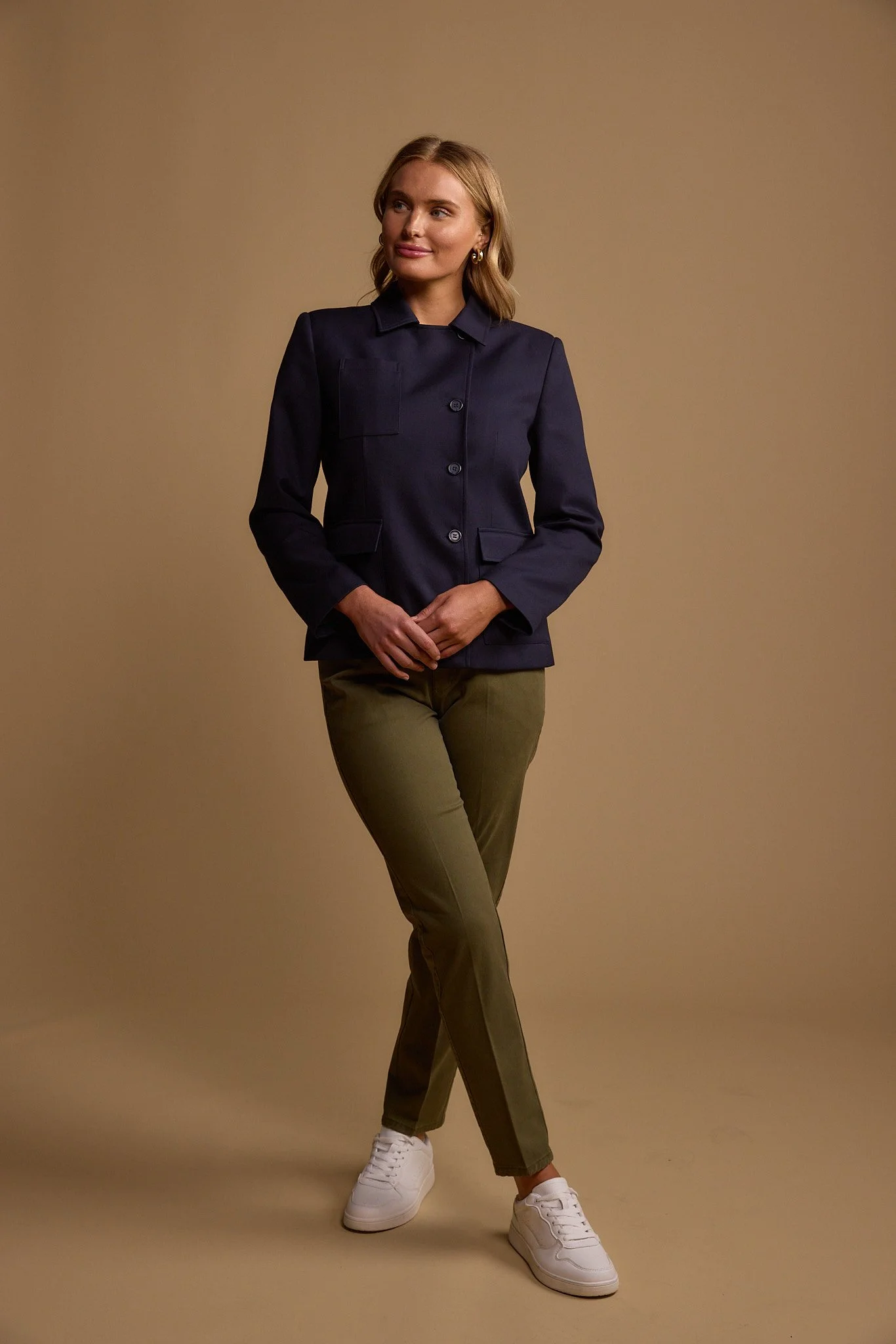 A woman wearing a dark blue blazer, olive green pants, and white sneakers, standing against a beige background.