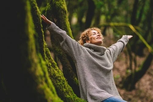 Person with curly hair sitting outdoors with arms outstretched, leaning against moss-covered trees