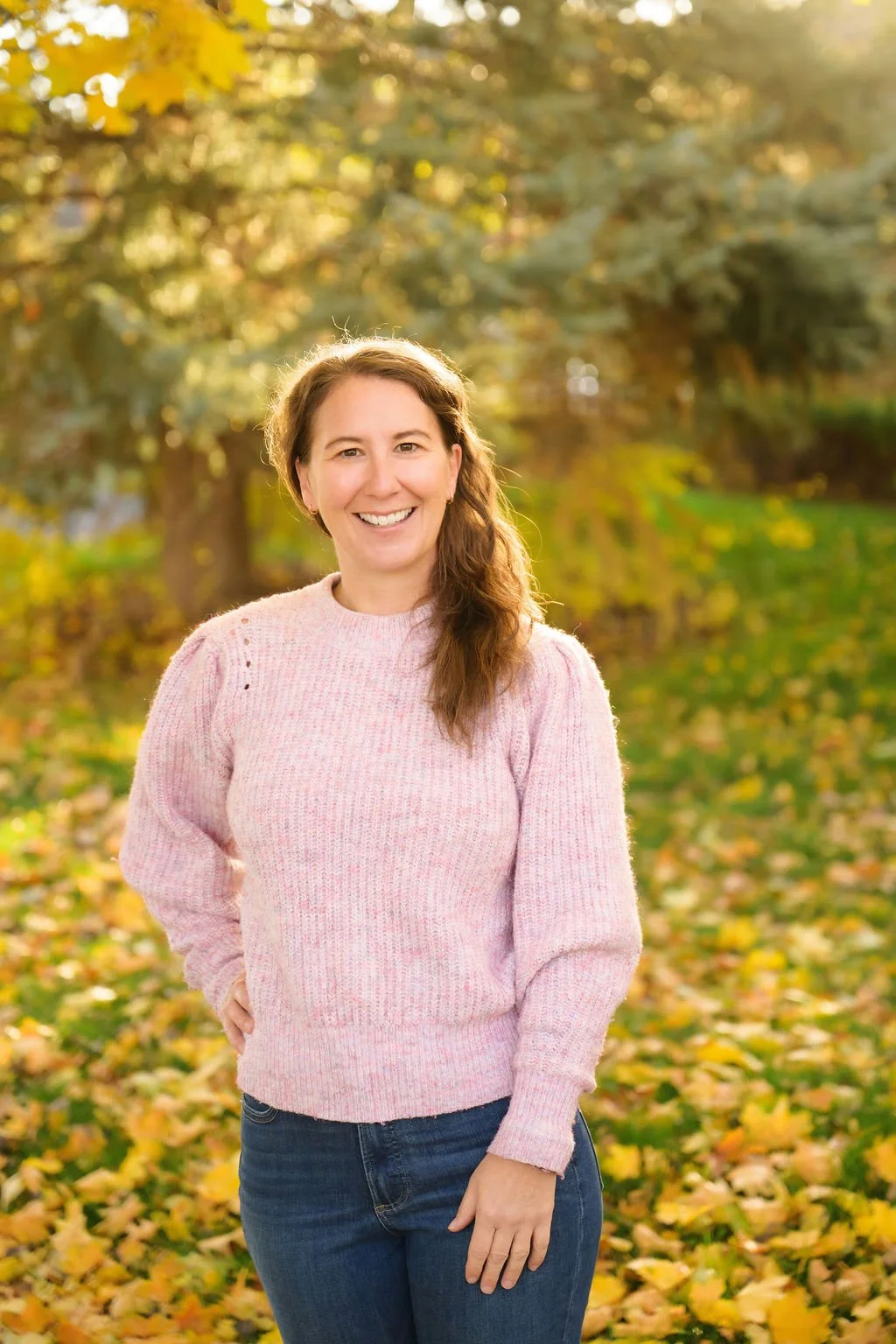 A smiling woman outdoors during autumn, wearing a pink sweater and jeans, standing in a park with fallen yellow leaves and trees in the background.