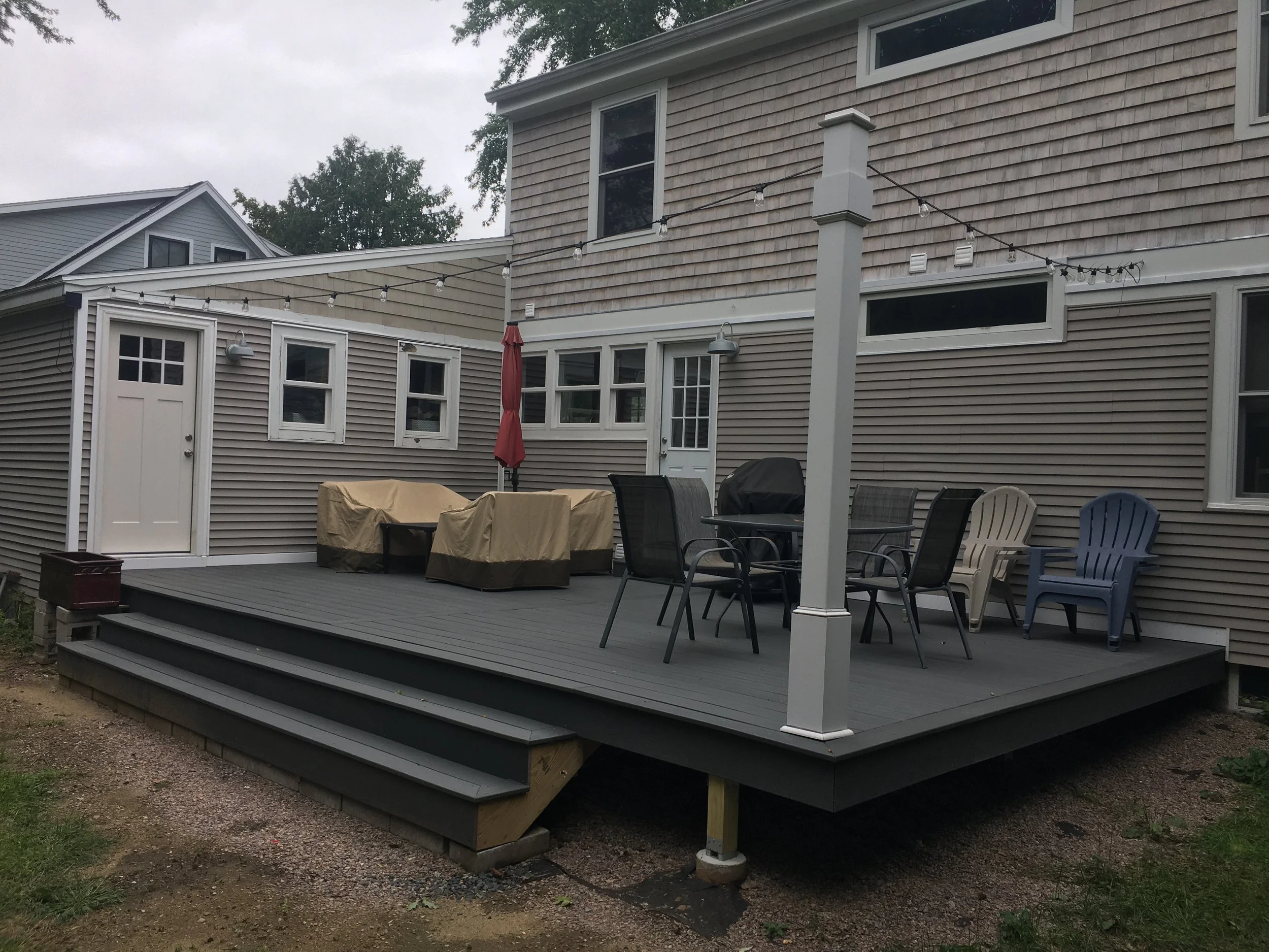 Back porch with outdoor furniture, including a table, chairs, and covered items, on a gray wooden deck with steps, attached to a house with beige siding, windows, and a door, under a cloudy sky.