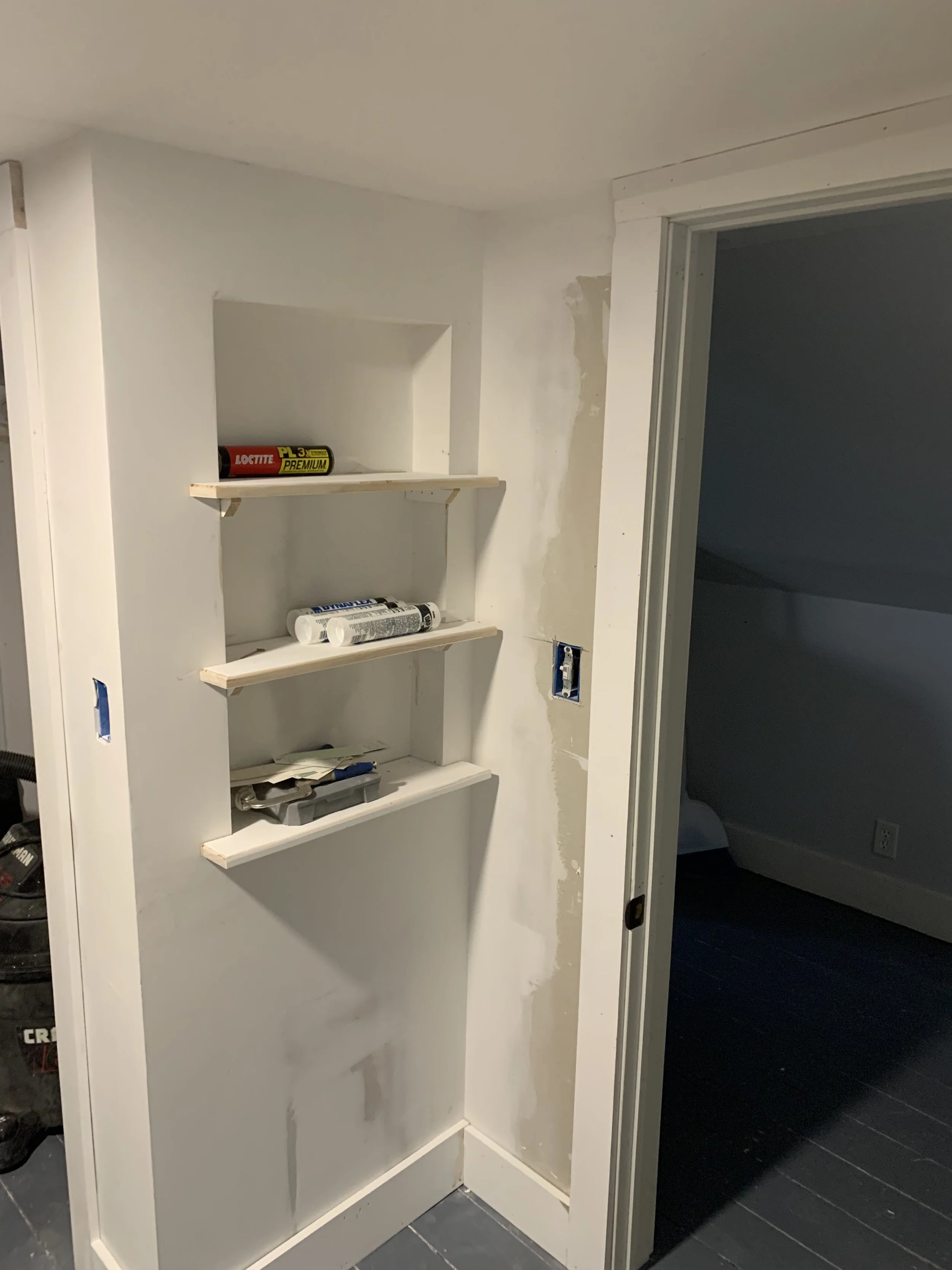 Built-in wall shelves with construction supplies in a partially finished room, next to an open doorway with no door installed.