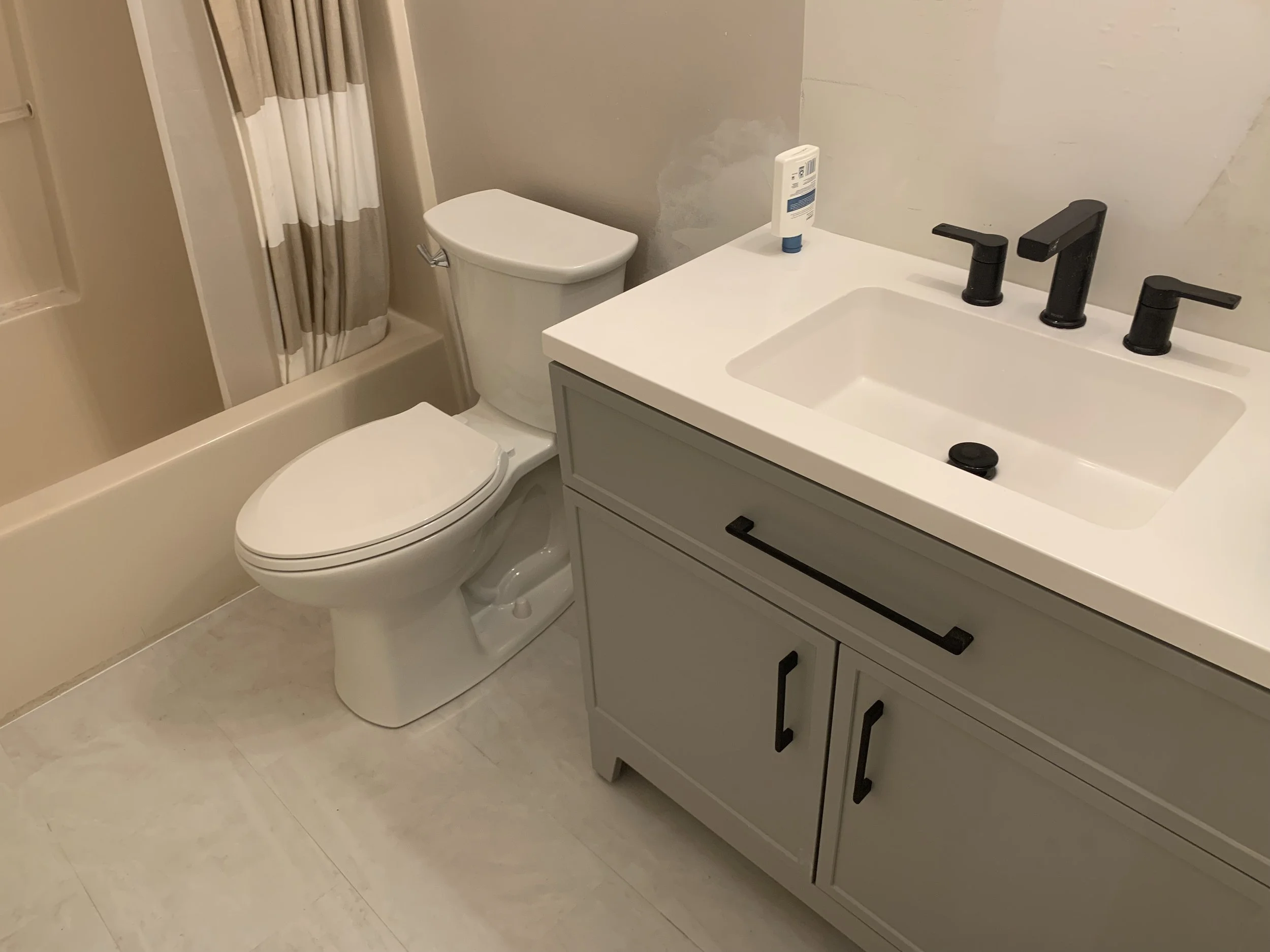 Bathroom with a toilet, a gray vanity with black handles, a white sink with black faucet fixtures, a bottle of lotion on the sink, and a shower curtain with beige and white stripes.