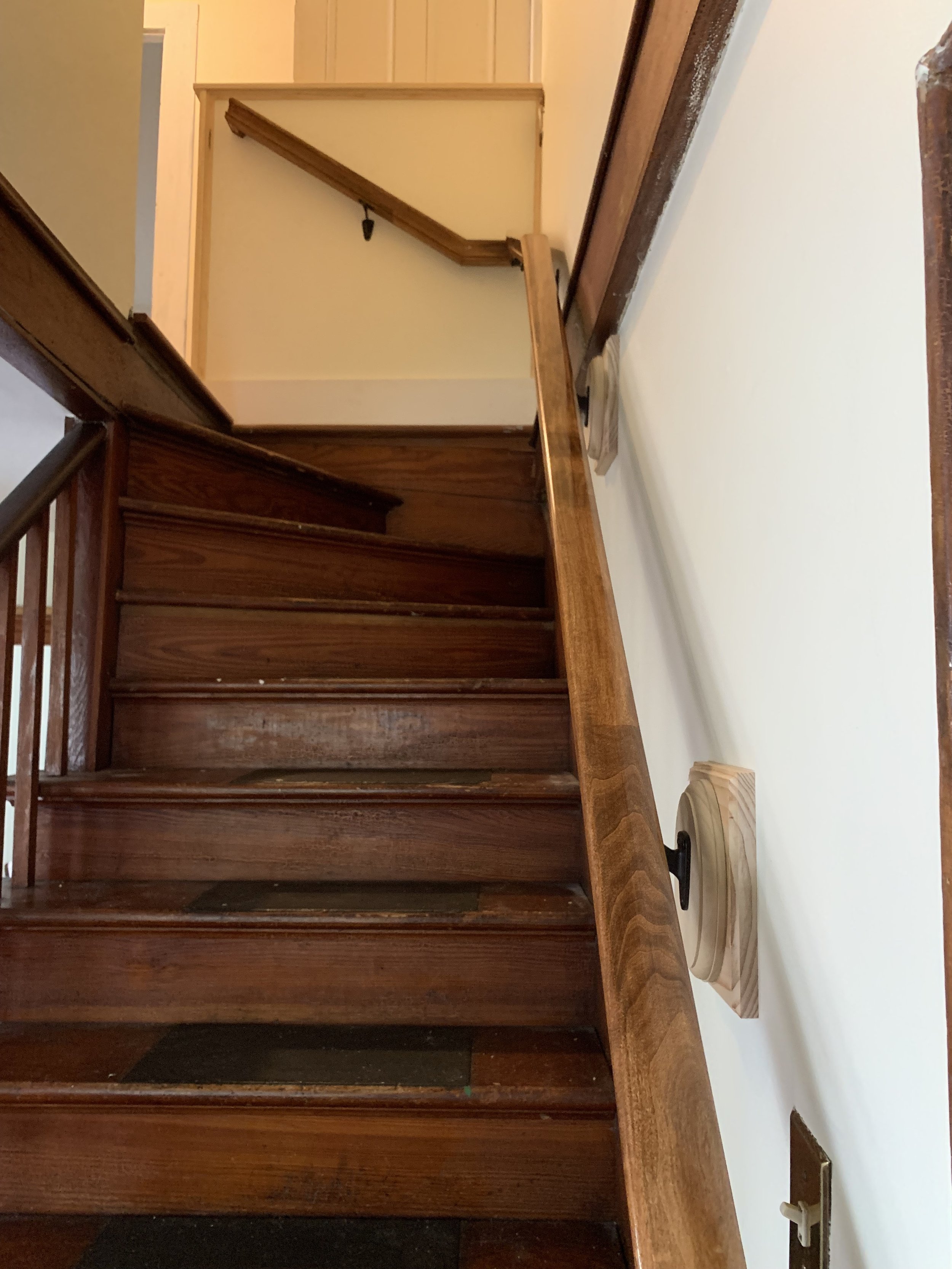 Wooden staircase with a handrail, featuring a newel post on the wall, leading up to a landing and a doorway.