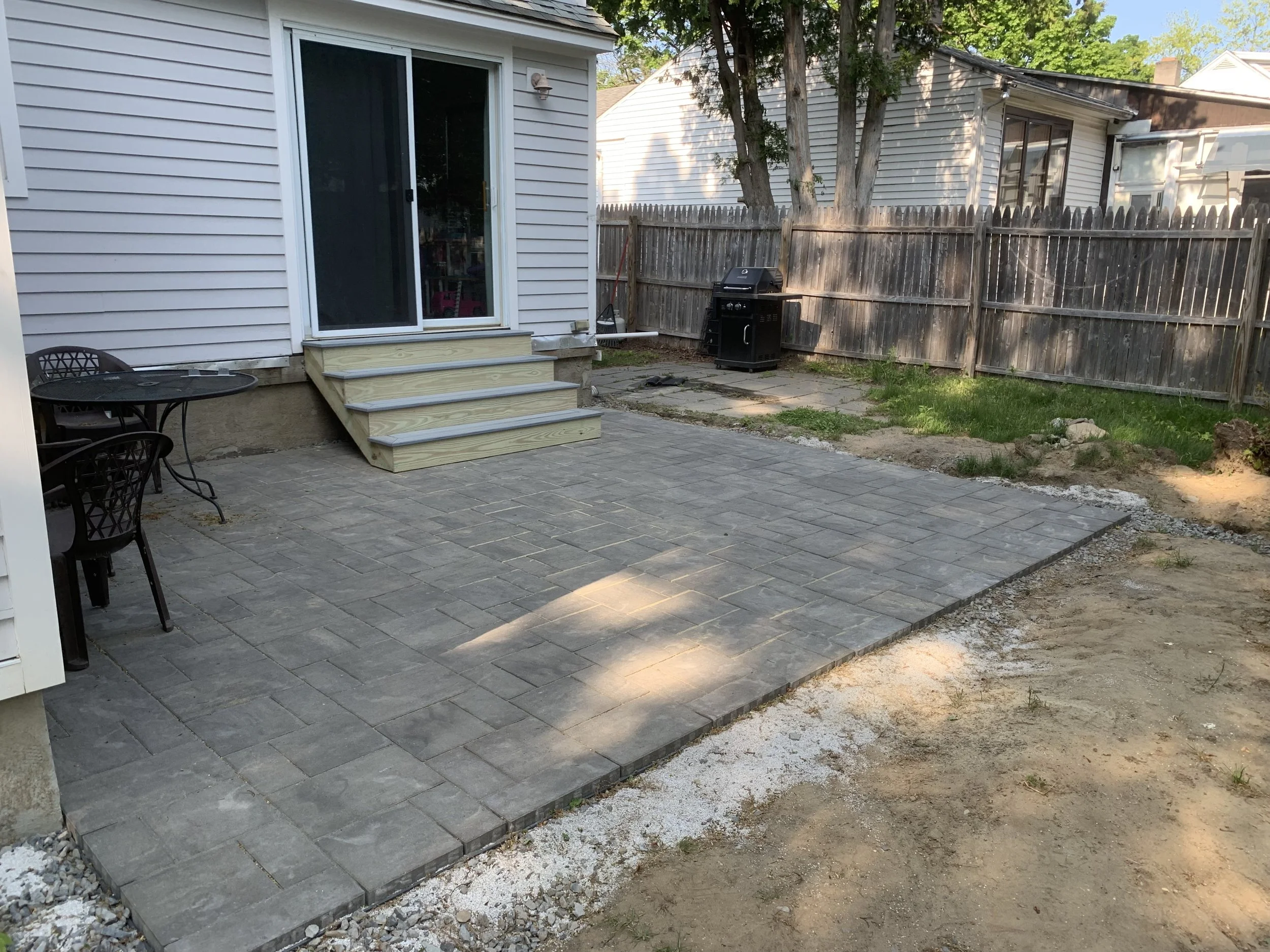 Backyard patio with new gray stone pavers, small staircase leading to a sliding glass door, outdoor dining table with chairs, and a black barbecue grill near a wooden fence.