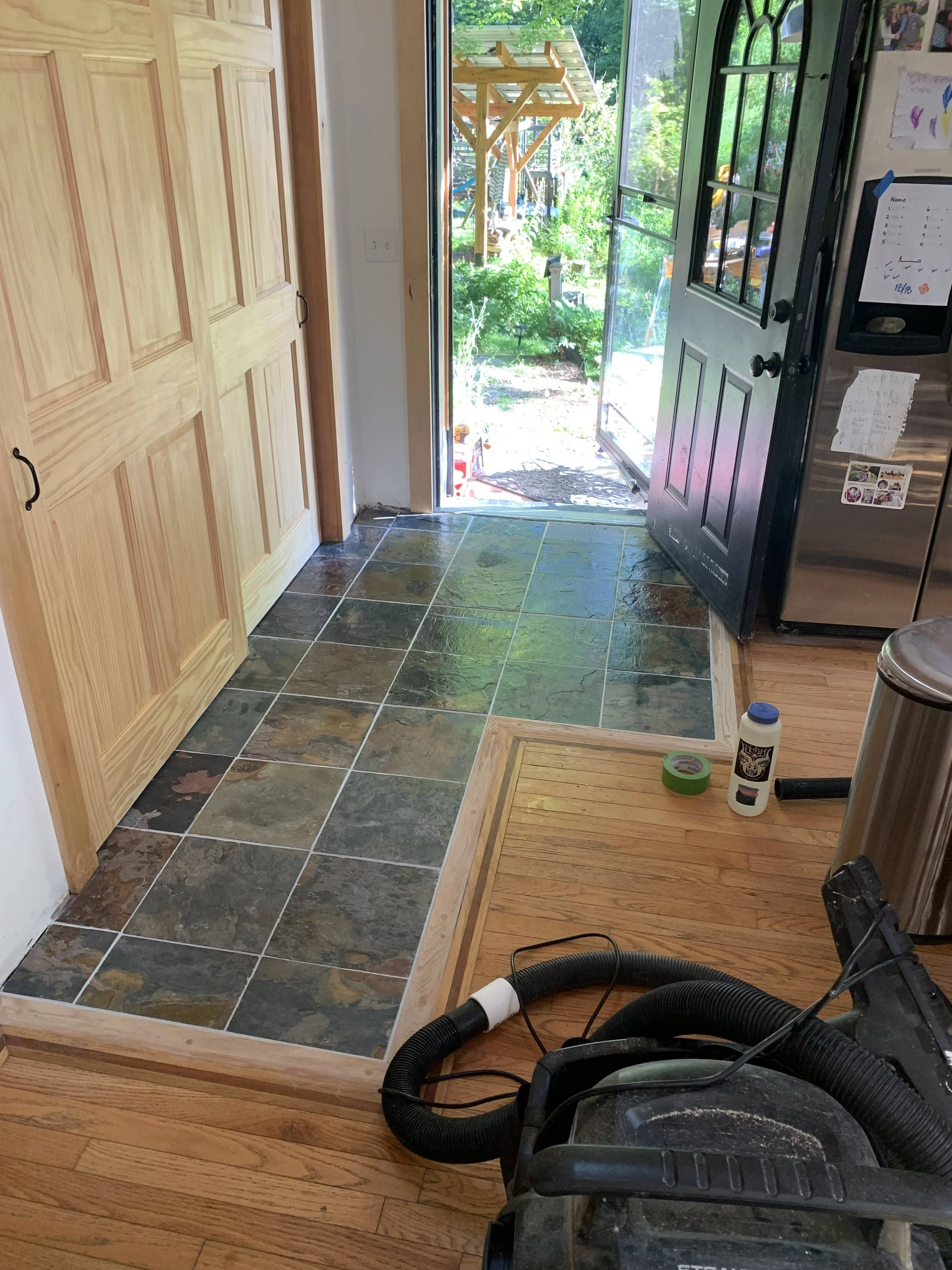 View of a partially renovated front door area with slate tile flooring, a black door, and a view outside of a garden with green plants and a wooden pergola.