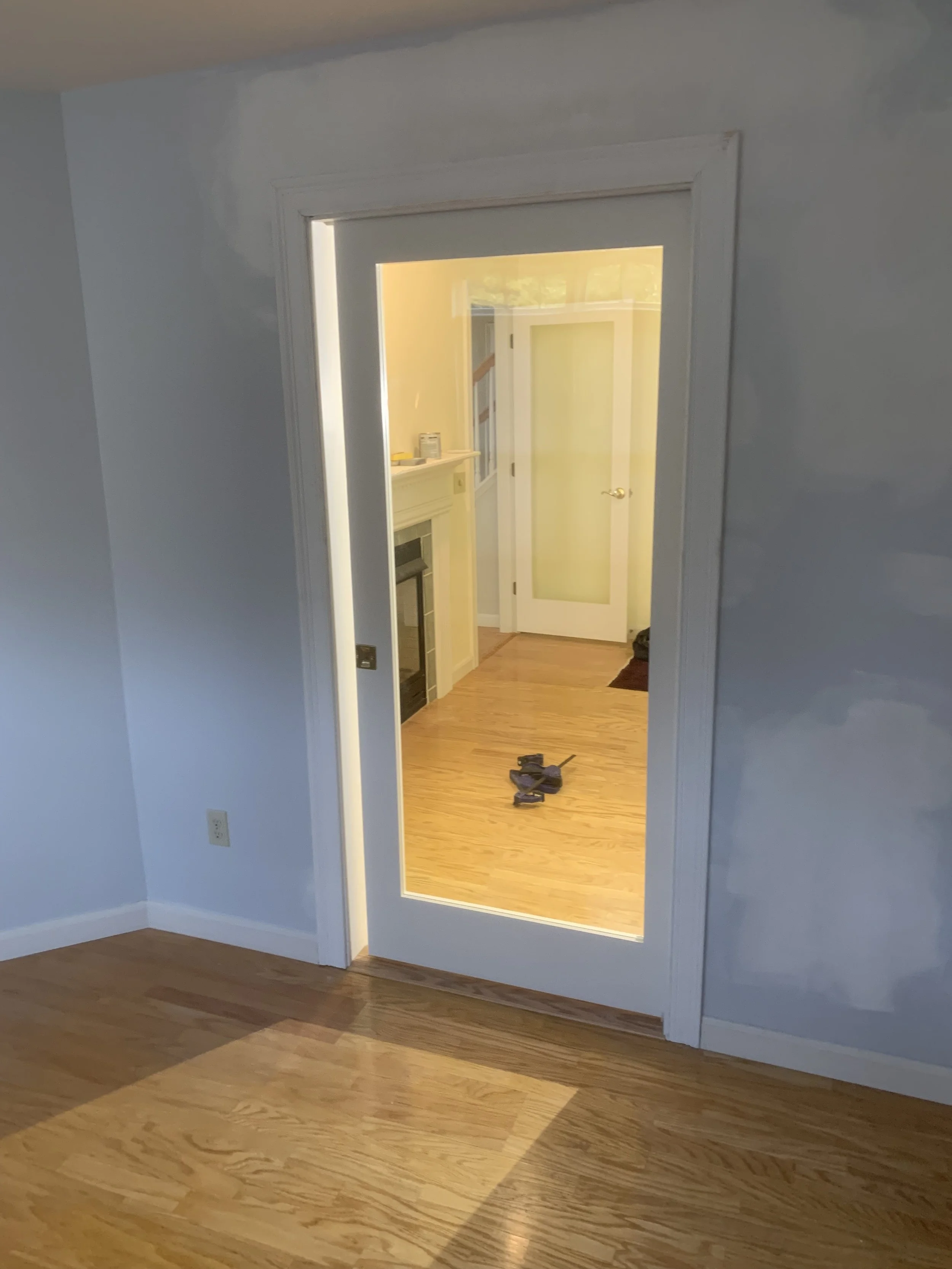 Interior view of a room with wooden floor, framing a door that opens into another room with wooden floor and a fireplace, with tools on the floor.