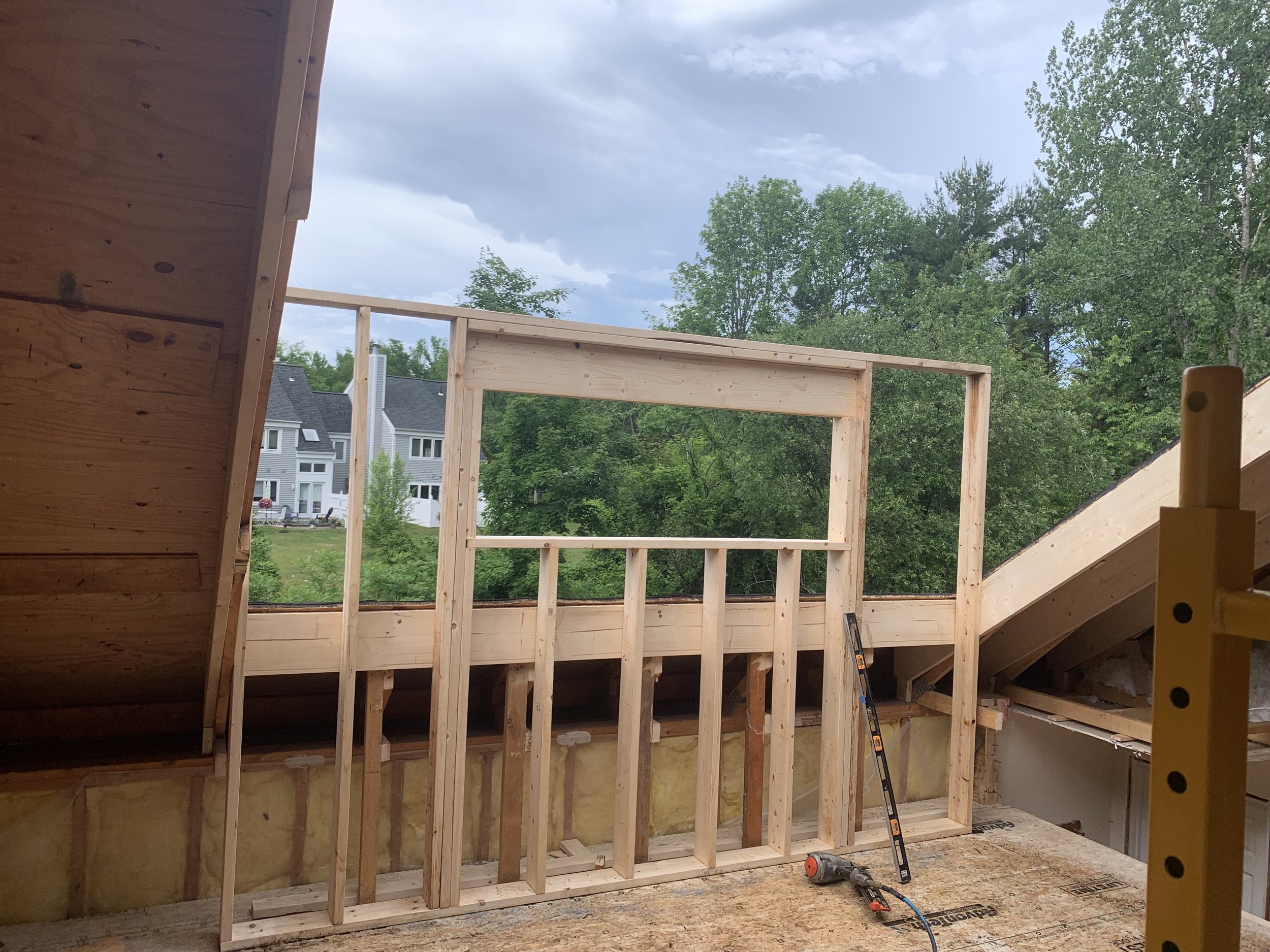 wooden framing in a construction site with an unfinished wall, a level tool, and a drill, with trees and houses in the background.