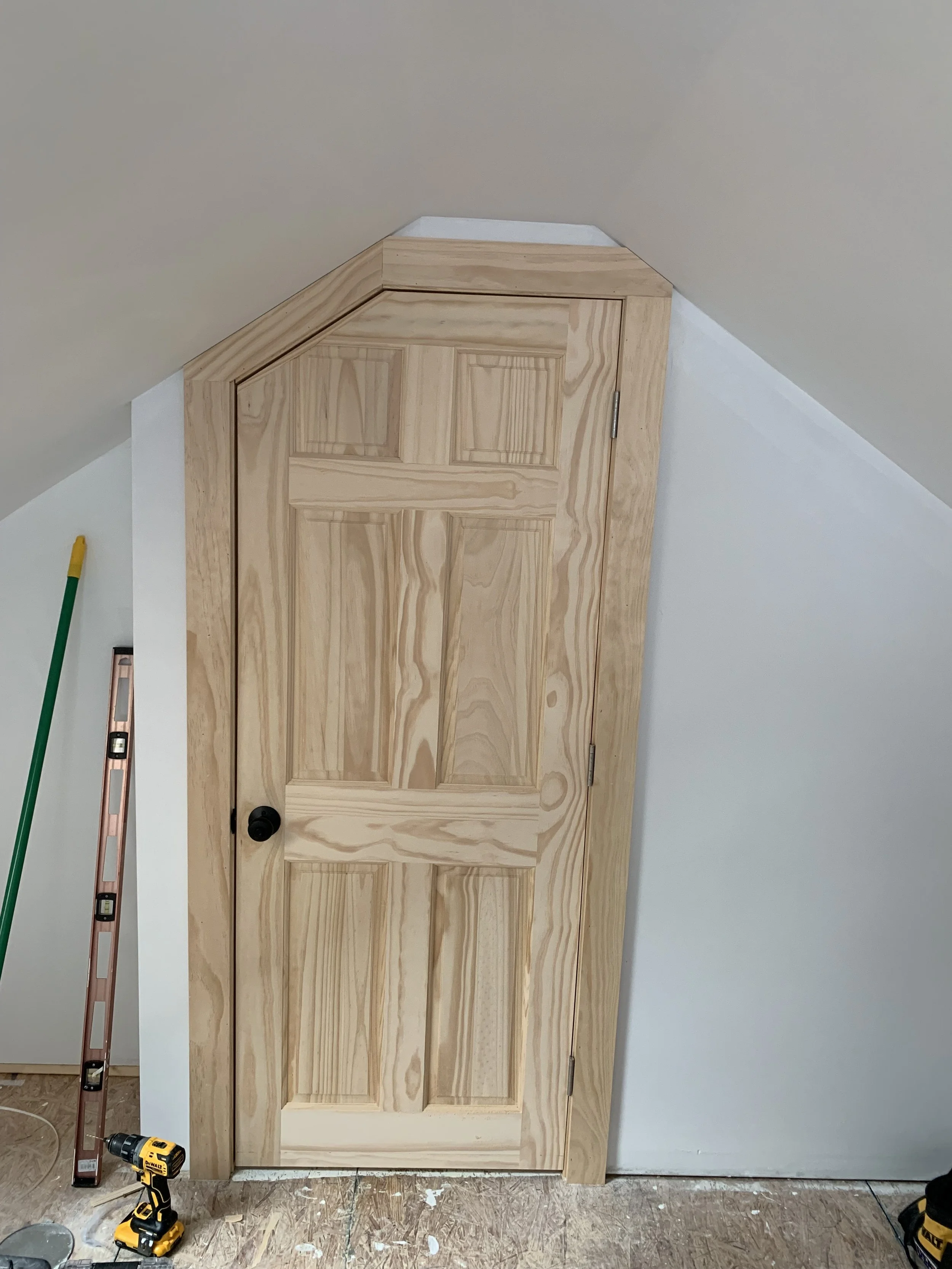 Wooden door installed under a sloped ceiling, with a black doorknob and visible hinges, in a room with construction tools nearby.