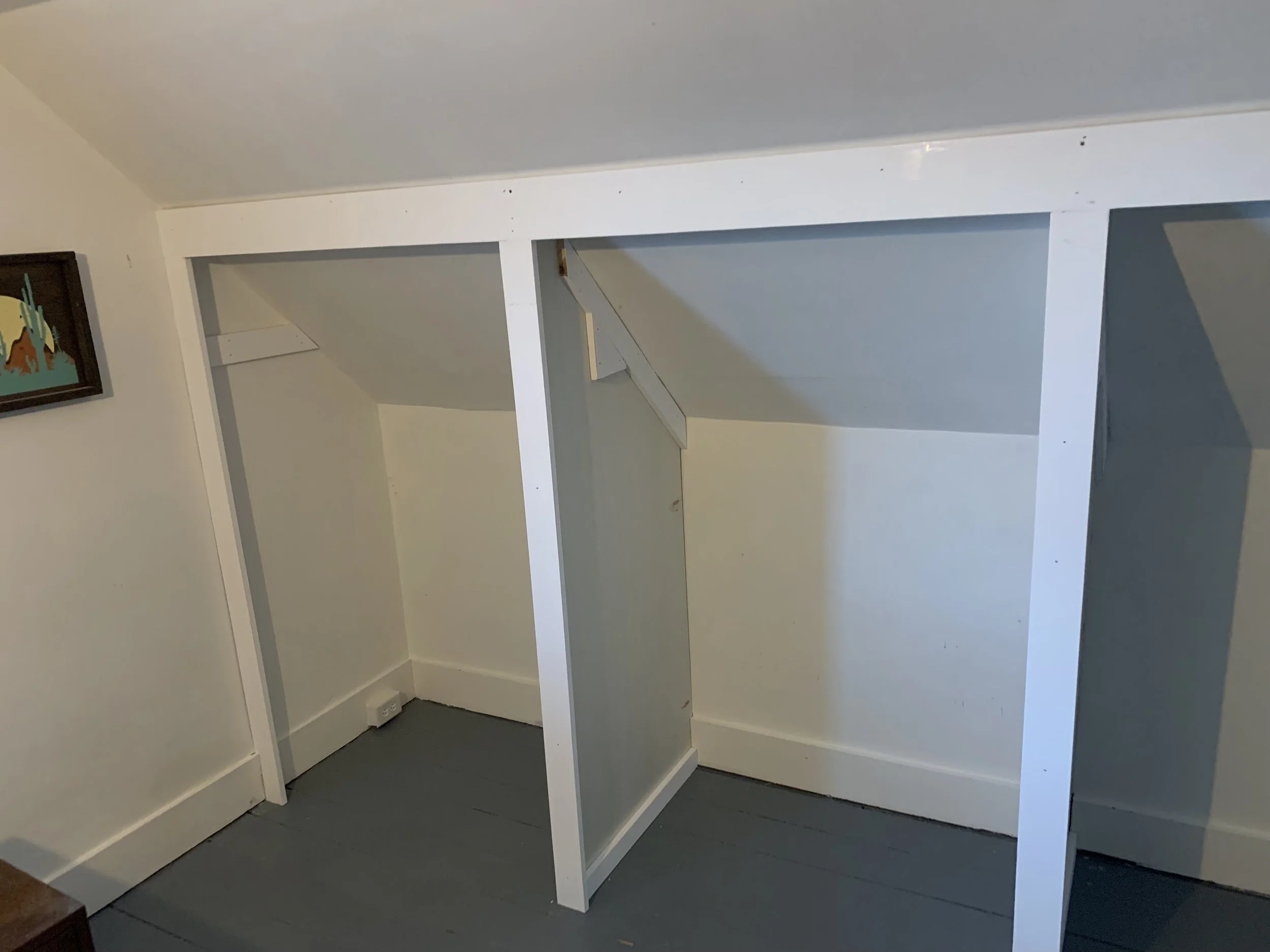 Empty built-in closet with three sections, painted white, located under a sloped ceiling in a room with light-colored walls and gray flooring. A small framed picture is on the wall to the left.