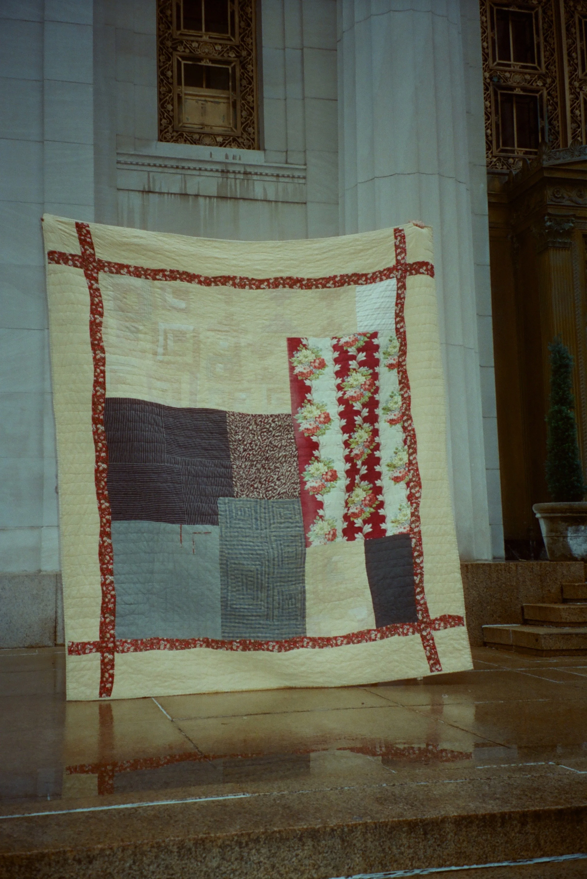 A quilt with a patchwork of various fabric patterns and colors, bordered in red floral fabric, displayed against an ornate building interior with marble walls and columns.