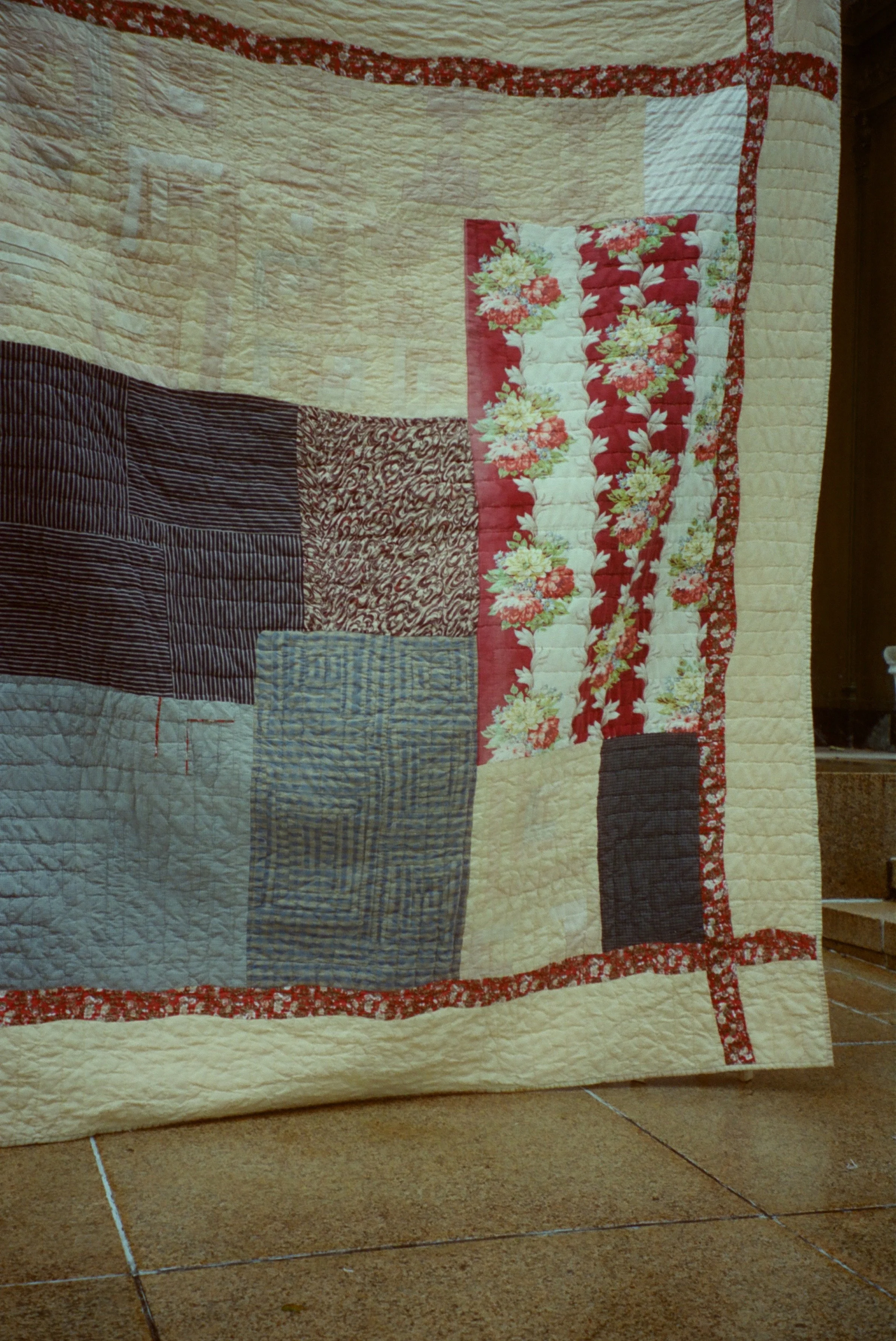 A quilt with various patterned fabric blocks, bordered with a floral red and white trim, lying on the floor.