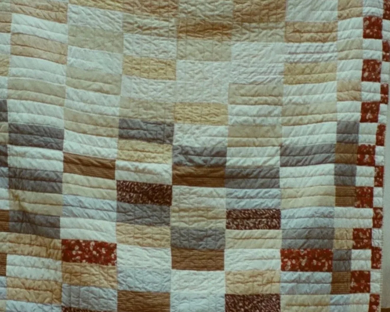 Close-up of a quilt with a pattern of rectangular fabric pieces in beige, brown, red, gray, and white colors.