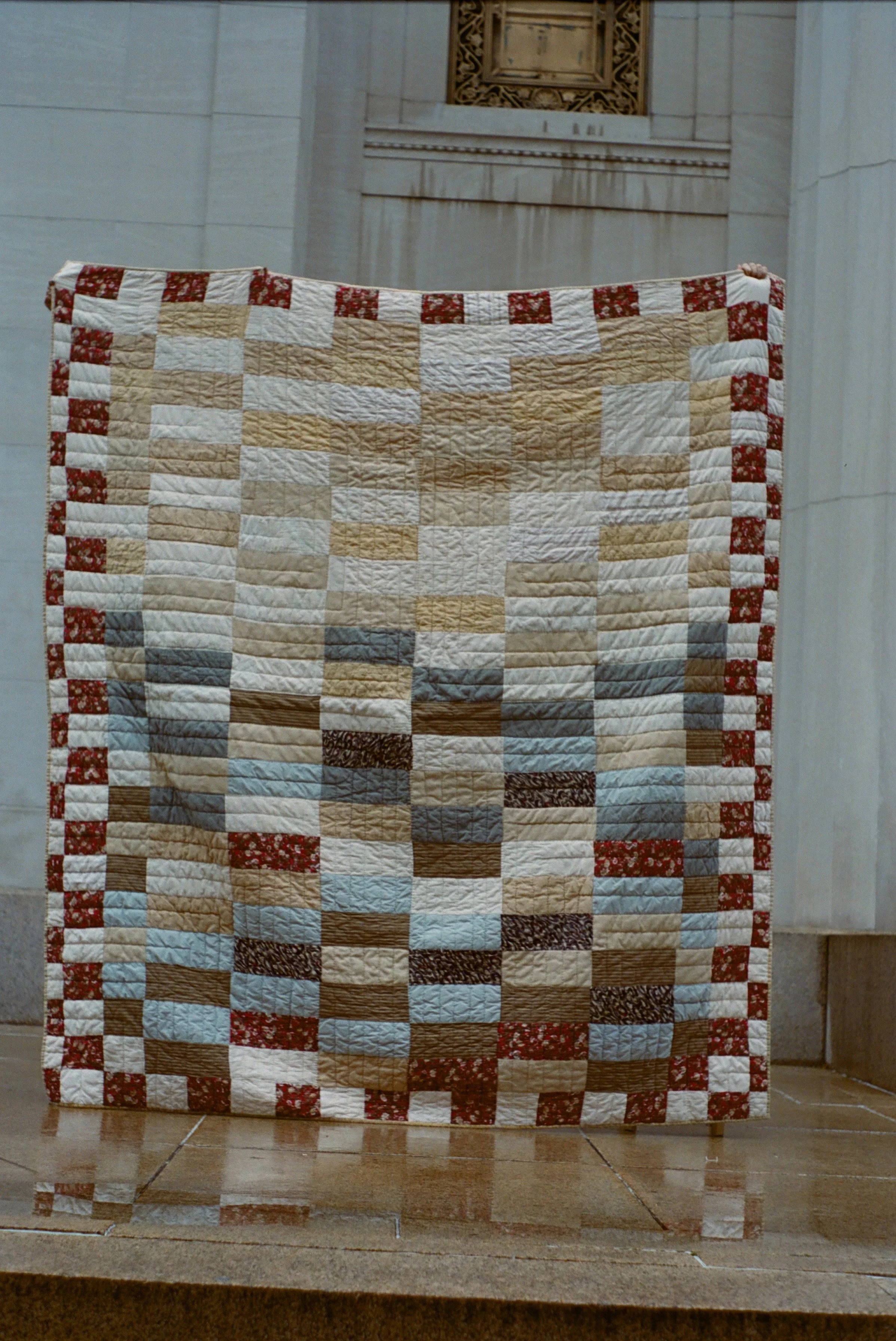 Person holding a quilt with a pixelated pattern in neutral and blue tones, framed with red and white floral fabric, inside a building with marble walls.