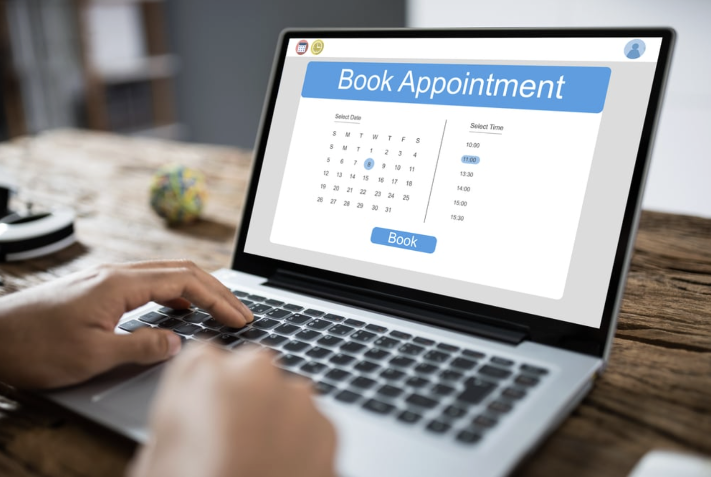 Laptop screen showing an online appointment booking webpage with a calendar, time selection, and a 'Book' button.