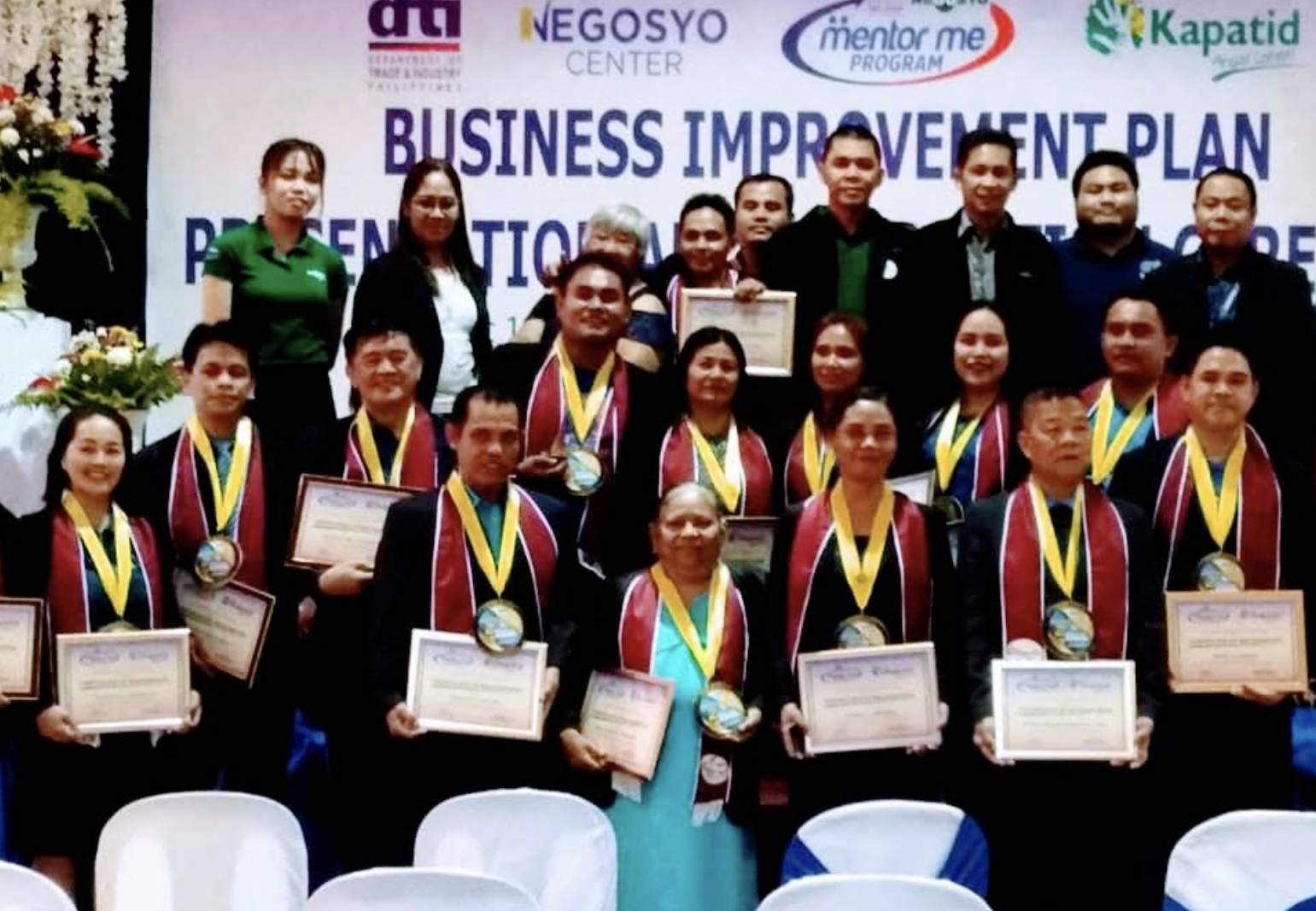 Group of people holding certificates and wearing medals at a business event or conference, with a backdrop displaying 'Business Improvement Plan' and other logos.