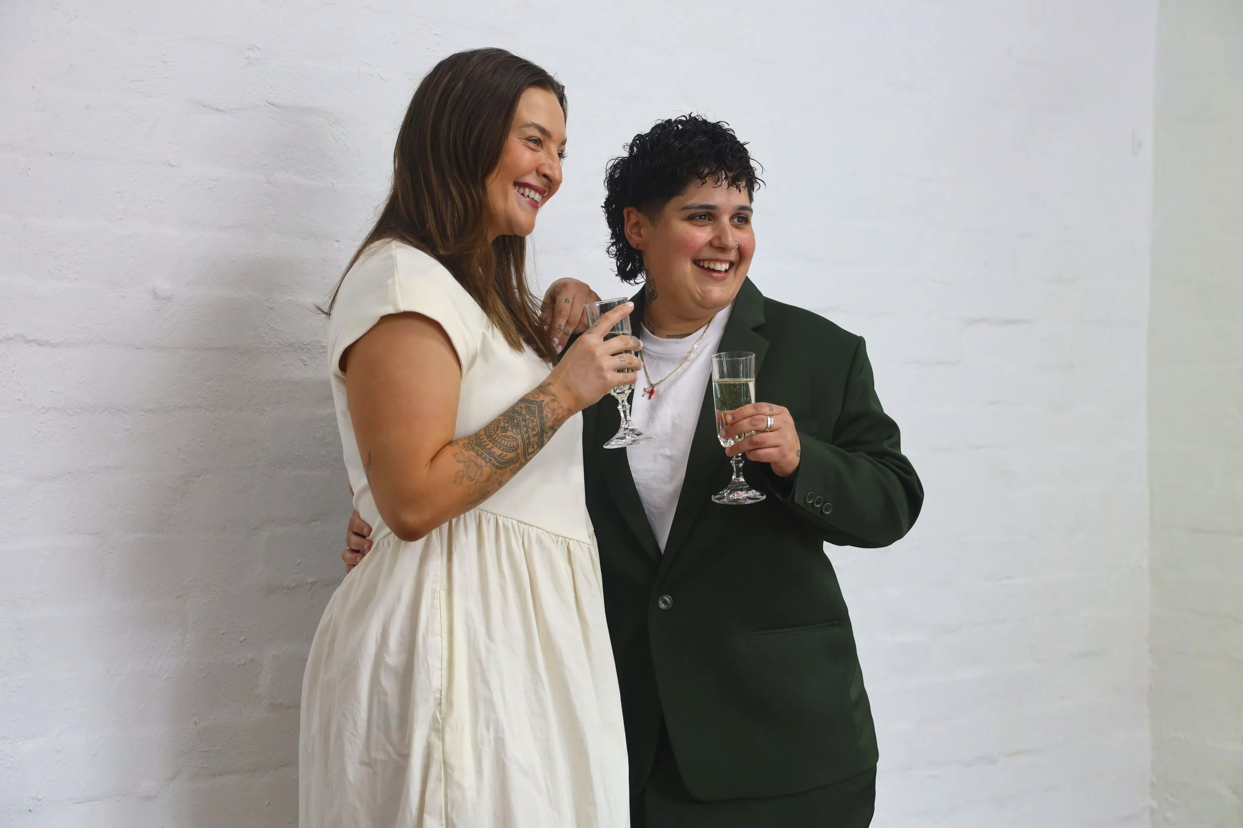 Two women smiling and holding glasses of champagne, standing against a white brick wall. One woman has long brown hair, tattoos on her arm, and is wearing a cream-colored dress. The other woman has short curly black hair, tattoos on her neck, and is dressed in a black blazer and white shirt.