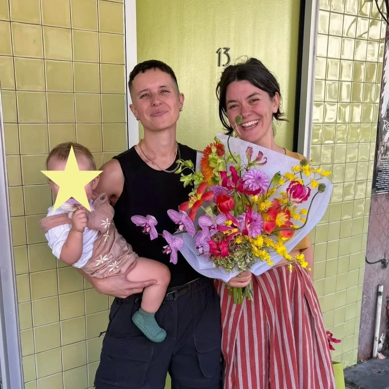 Jen the celebrant stands with their wife and baby on a street in Sydney. Jen's wife holds a large bouquet of colourful flowers. They are all smiling.
