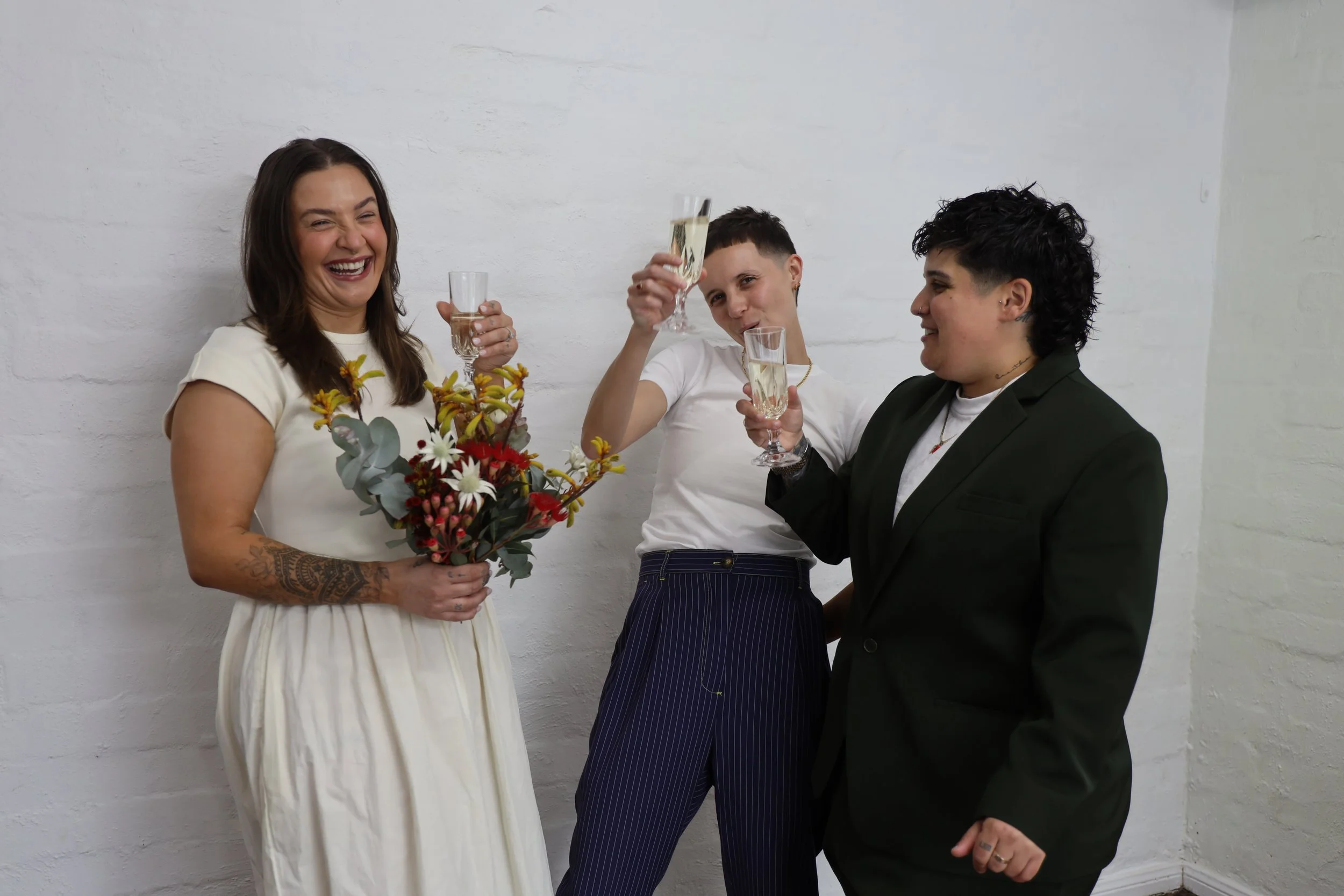 Jen the celebrant stands between two people who are getting married. They each raise a champagne glass in celebration, and are smiling.