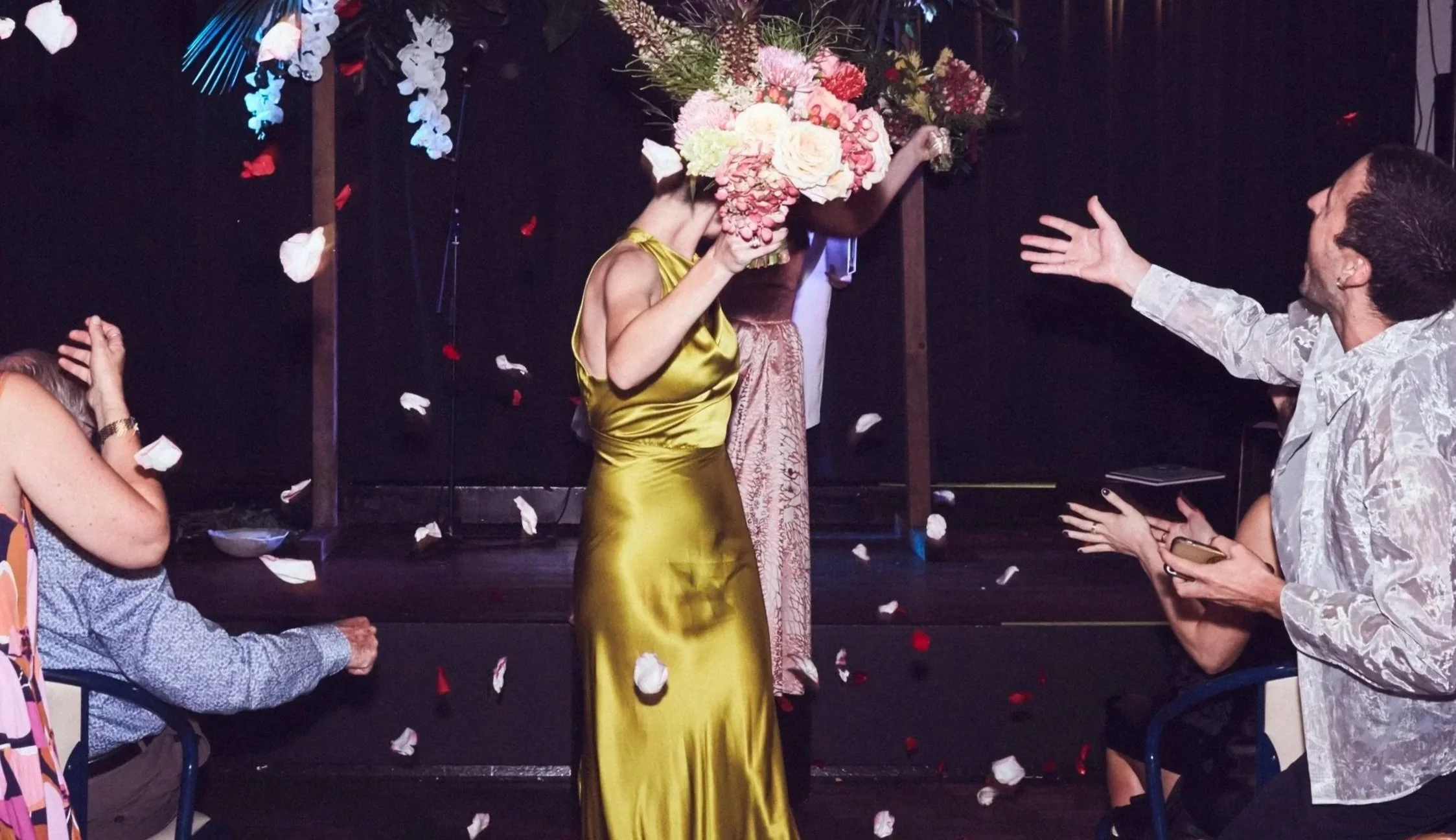 Two people walk down the aisle after getting married, holding a large bouquet of colourful flowers. A happy crowd throws flower petals on them as they walk.