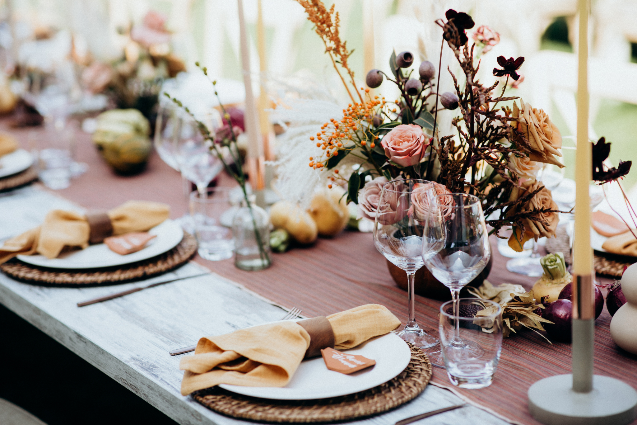 A table set for a dinner party with woven placemats, white plates, beige napkins with brown napkin rings, and clear wine glasses. The table has a centerpiece with pink roses, brown and black foliage, small yellow and purple decorative items, and scattered fruits and vegetables. Yellow candles are also part of the decor.