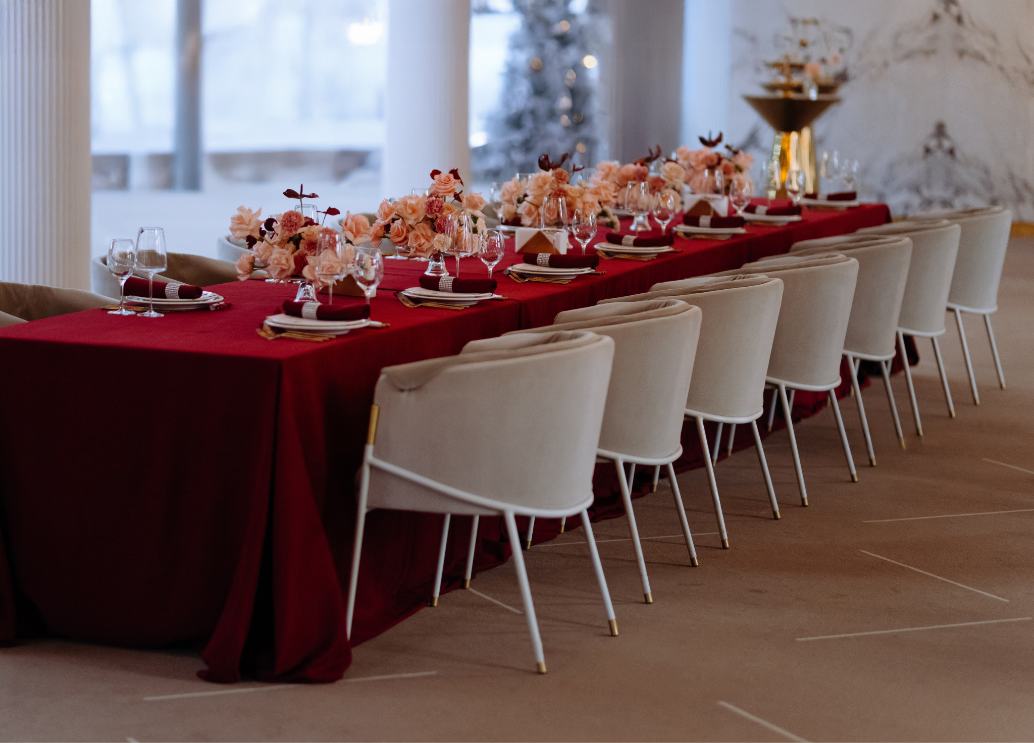 Elegant banquet table decorated with pink flowers, wine glasses, and red and white napkins in a sophisticated event space with a Christmas tree in the background. Social event, themed dinner with friends and family, girls' night, bridal events