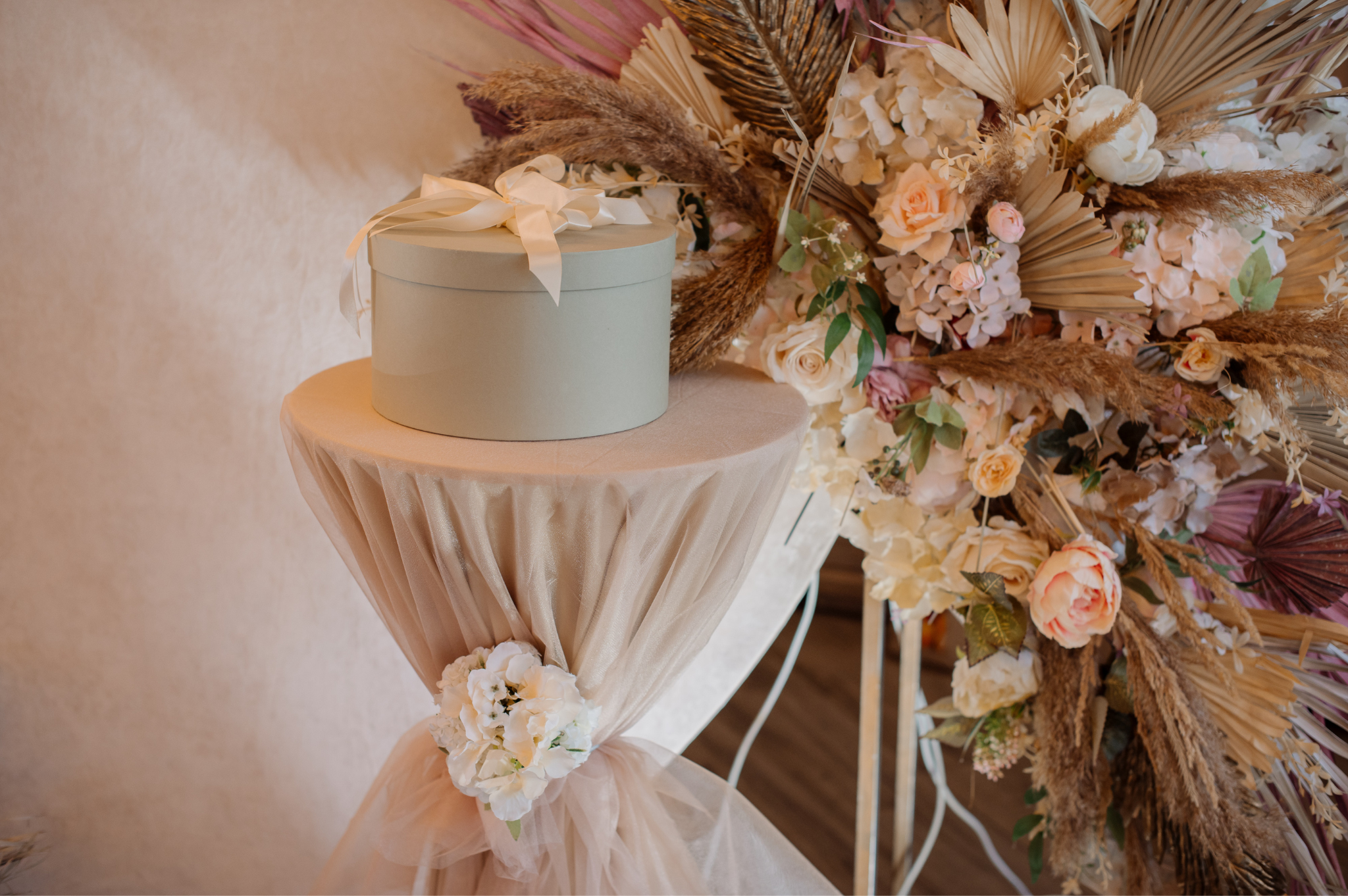 A round gift box with a ribbon bow on top, placed on a small table draped with pink fabric and floral decorations, near a large floral arrangement of white and pink flowers and dried plants.