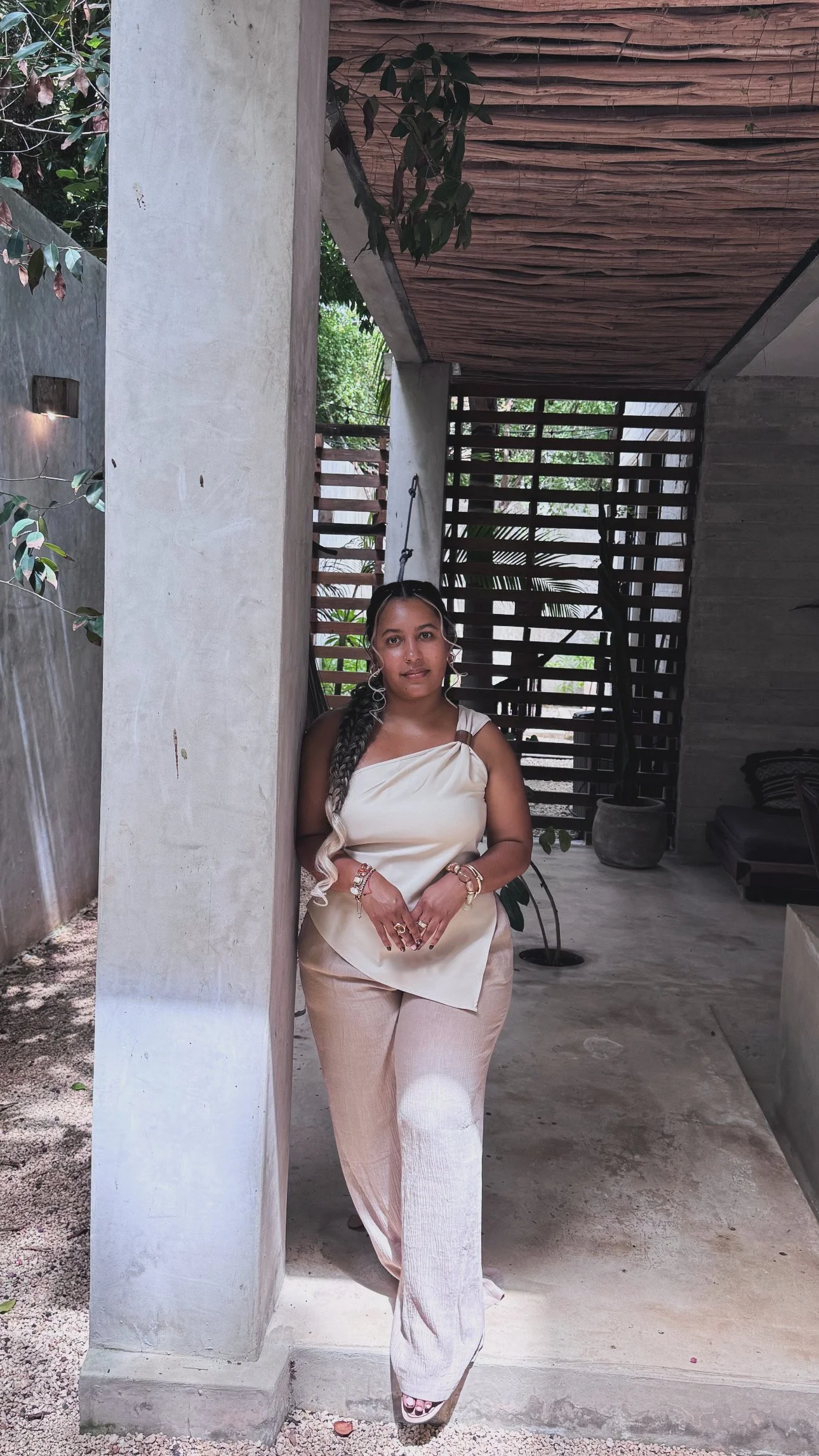 A woman event planner with long, braided hair and jewelry, wearing a cream-colored top and beige pants, standing outdoors next to a concrete wall and hanging from a swing in a modern, minimalist patio area.