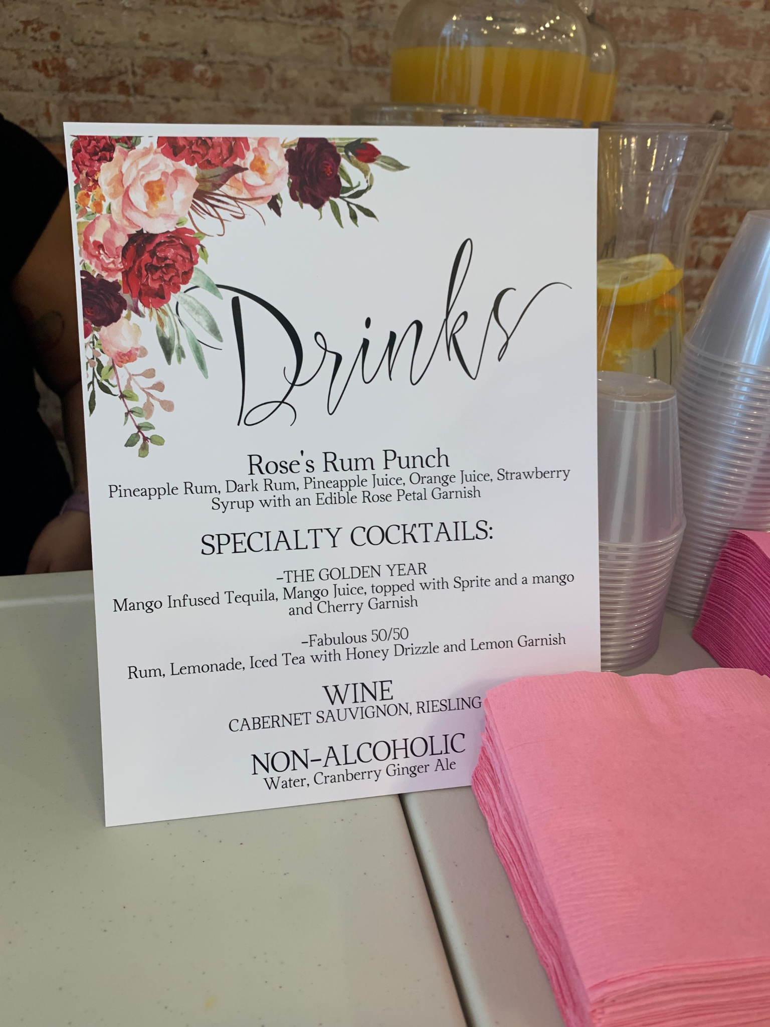 A customized/ personalized party menu board with floral decorations listing drinks including specialty cocktails, wine, and non-alcoholic options, placed on a table with pink napkins and drink dispensers in the background. birthday party, anniversary