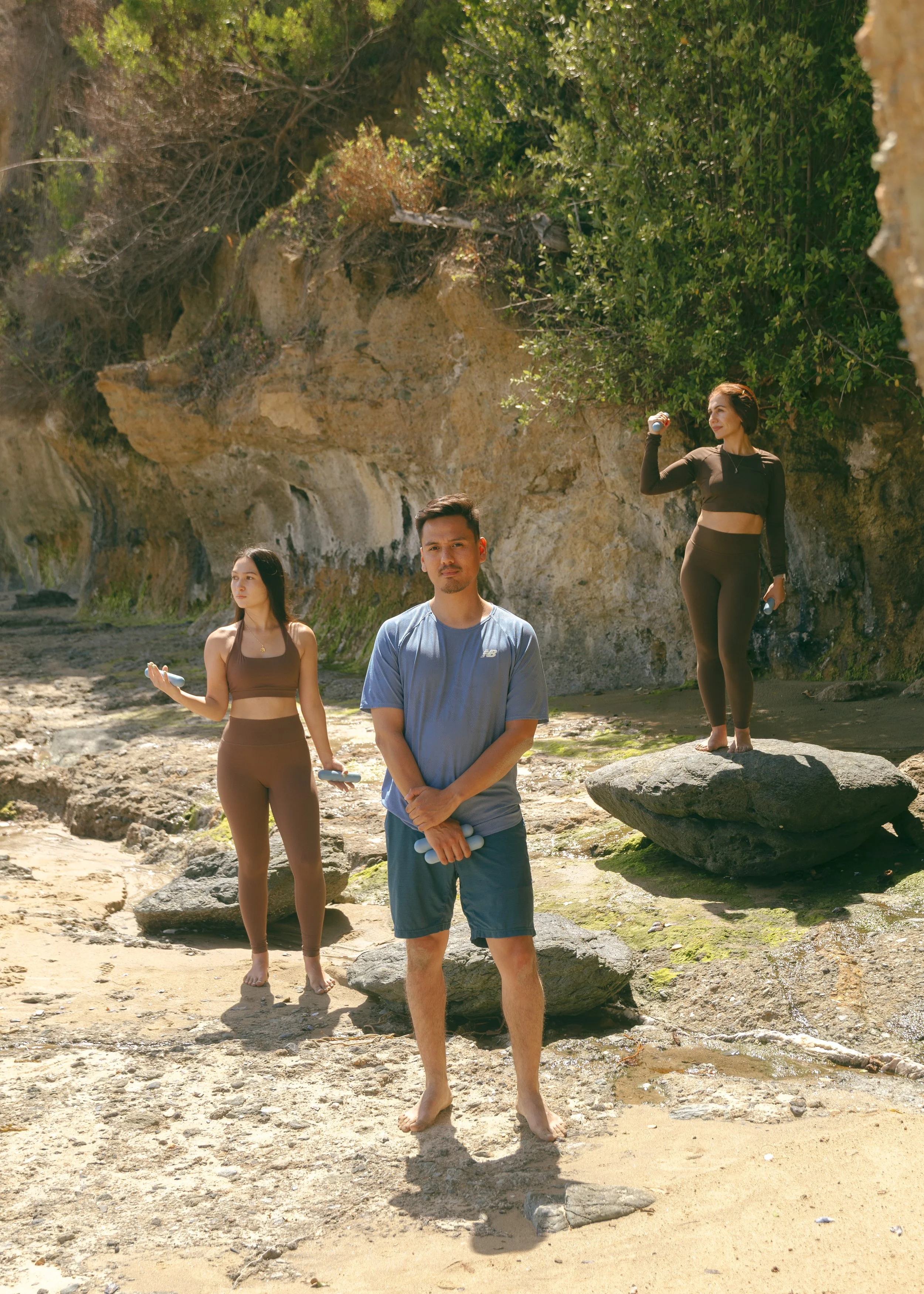 Three people (two women and a male) standing in various positions holding Bala weights on the beach.