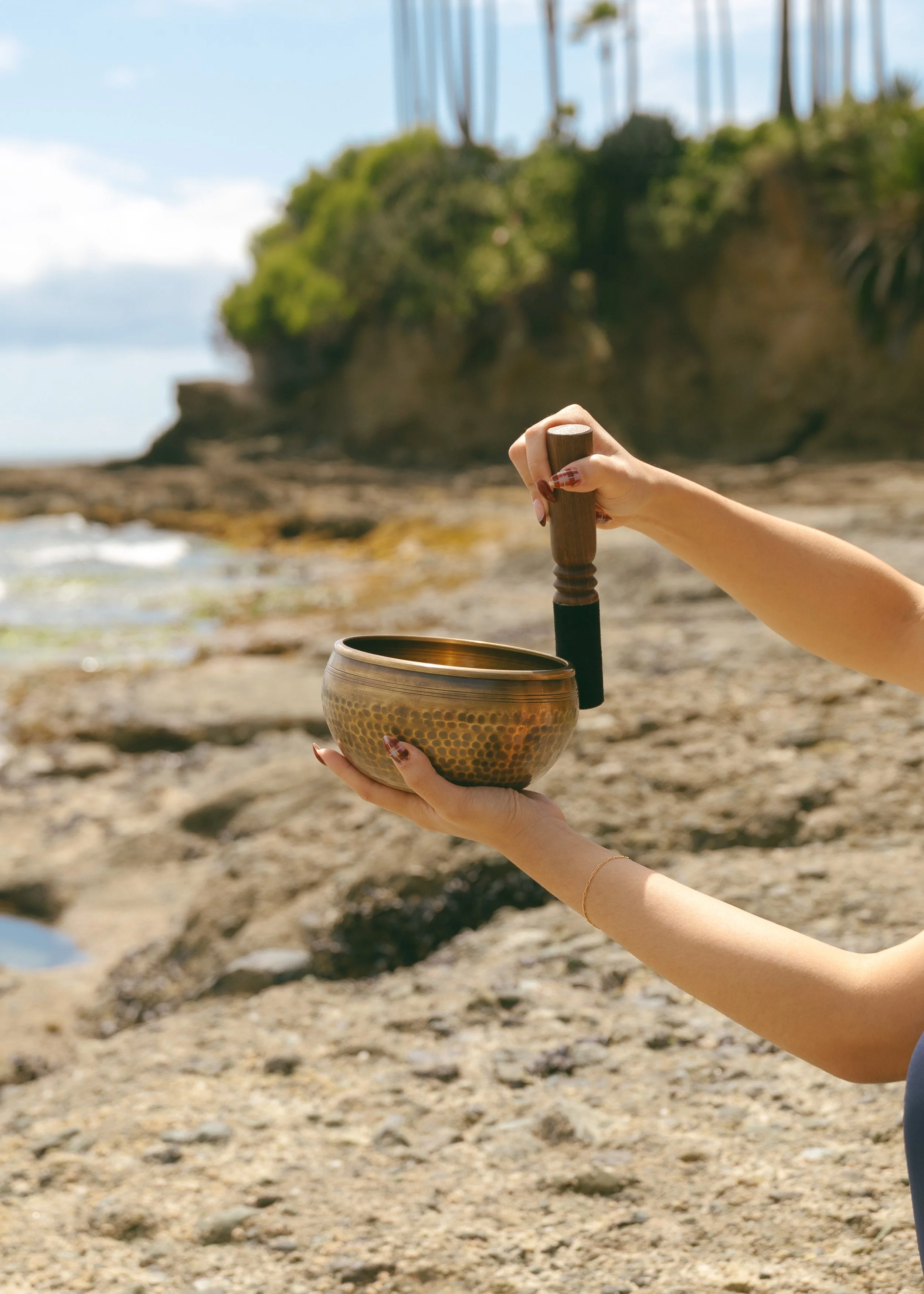 Arms playing a Tibetan sound bowl with the beach as a backdrop.